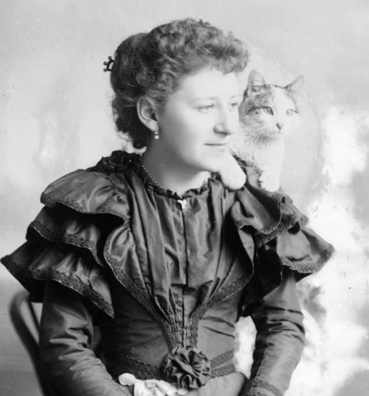 Black and white photo portrait of young white woman in a dark, ruffled Victorian dress. She is seated and looking happily off to the right of the viewer. On her left shoulder sits an adorable petite, shorthaired white and tabby cat which is also looking to the right of the viewer.