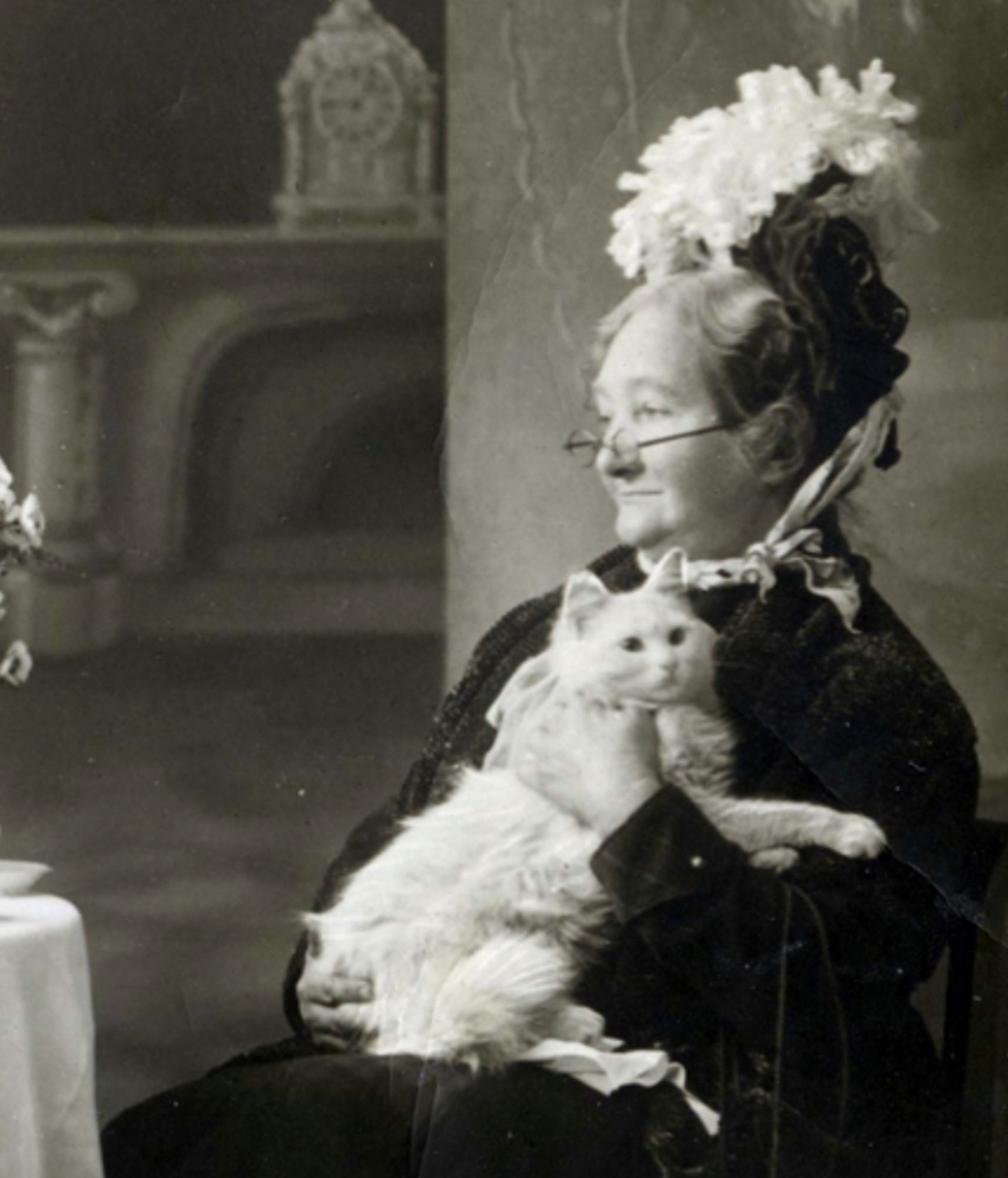 Black and white photo of an older white woman wearing a dark dress and a large hat festooned with an explosion of white flowers. She is sitting in a wooden chair at a table covered in a white tale cloth in a scene that seems to be arranged in a studio setting. In her arms she cradles a medium haired white cat.