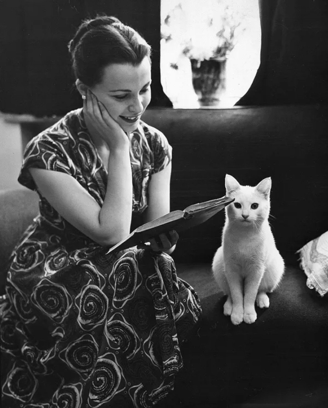 Black and white photo of a pretty, smiling young white woman in a boldly patterned shortsleeved dress, sitting on a couch reading an open book on her knee. Next to her sits a beautiful little white shorthaired cat that sit up looking curiously beyond the camera like it's about to dash off and chase something.