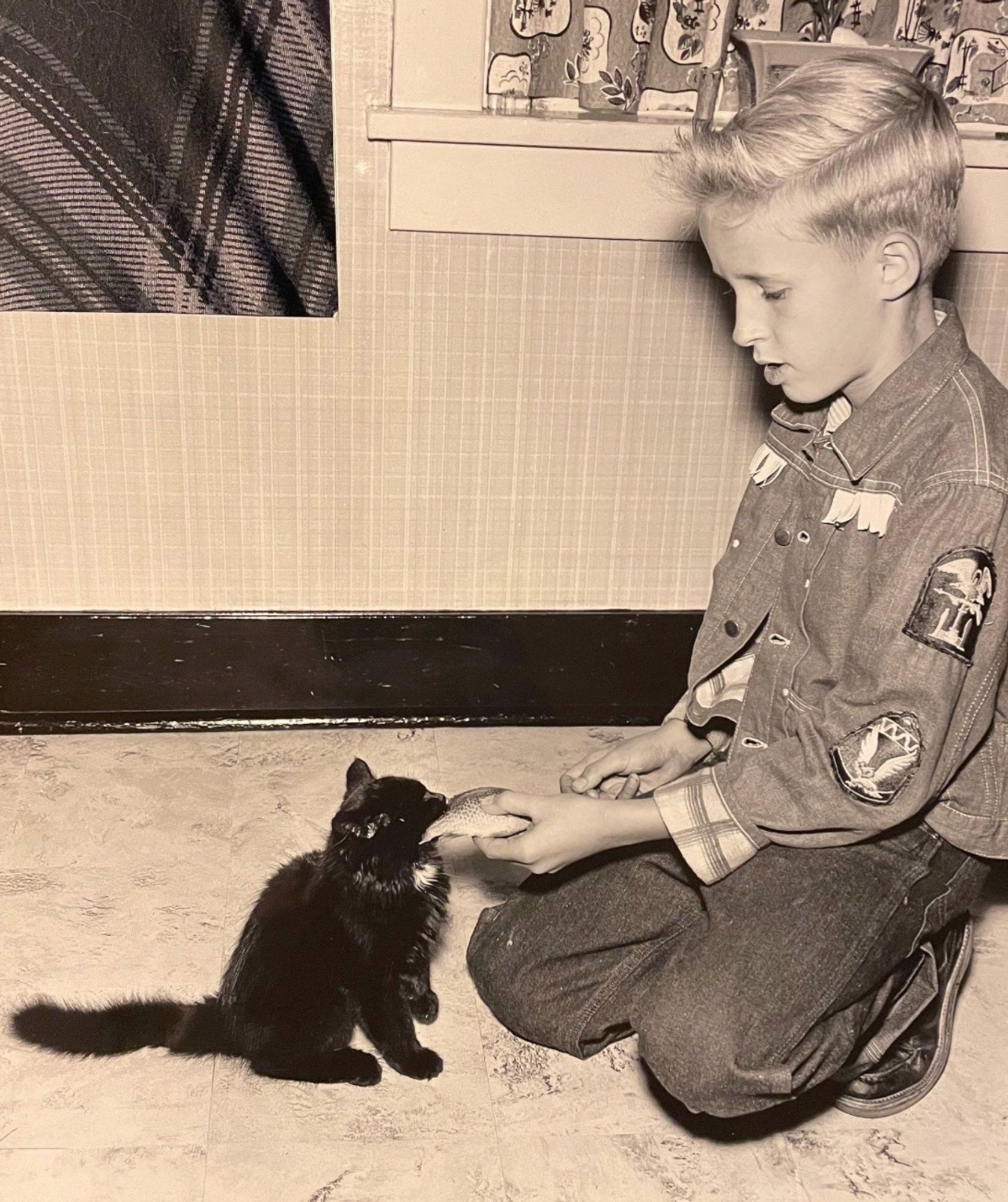 Black and white photo of a young white boy kneeling on a linoleum floor, offer a small, whole fish to a fluffy black kitten with a white spot at its throat. A square of the photo is missing in the upper left corner and you can see my grey plaid pants underneath.