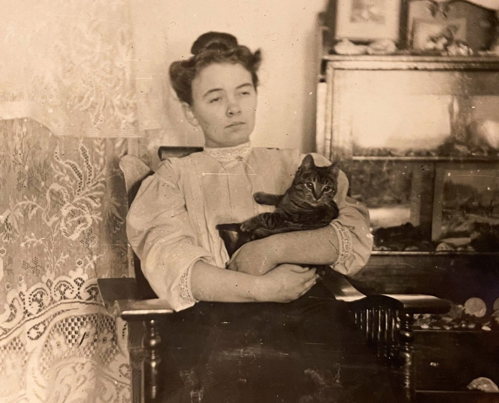 A sepia toned photo of a woman with a stern, skeptical expression on her face. She’s wearing early 20th century clothing, sitting in a wooden chair, and lovingly cradling a dark tabby cat in her arms.