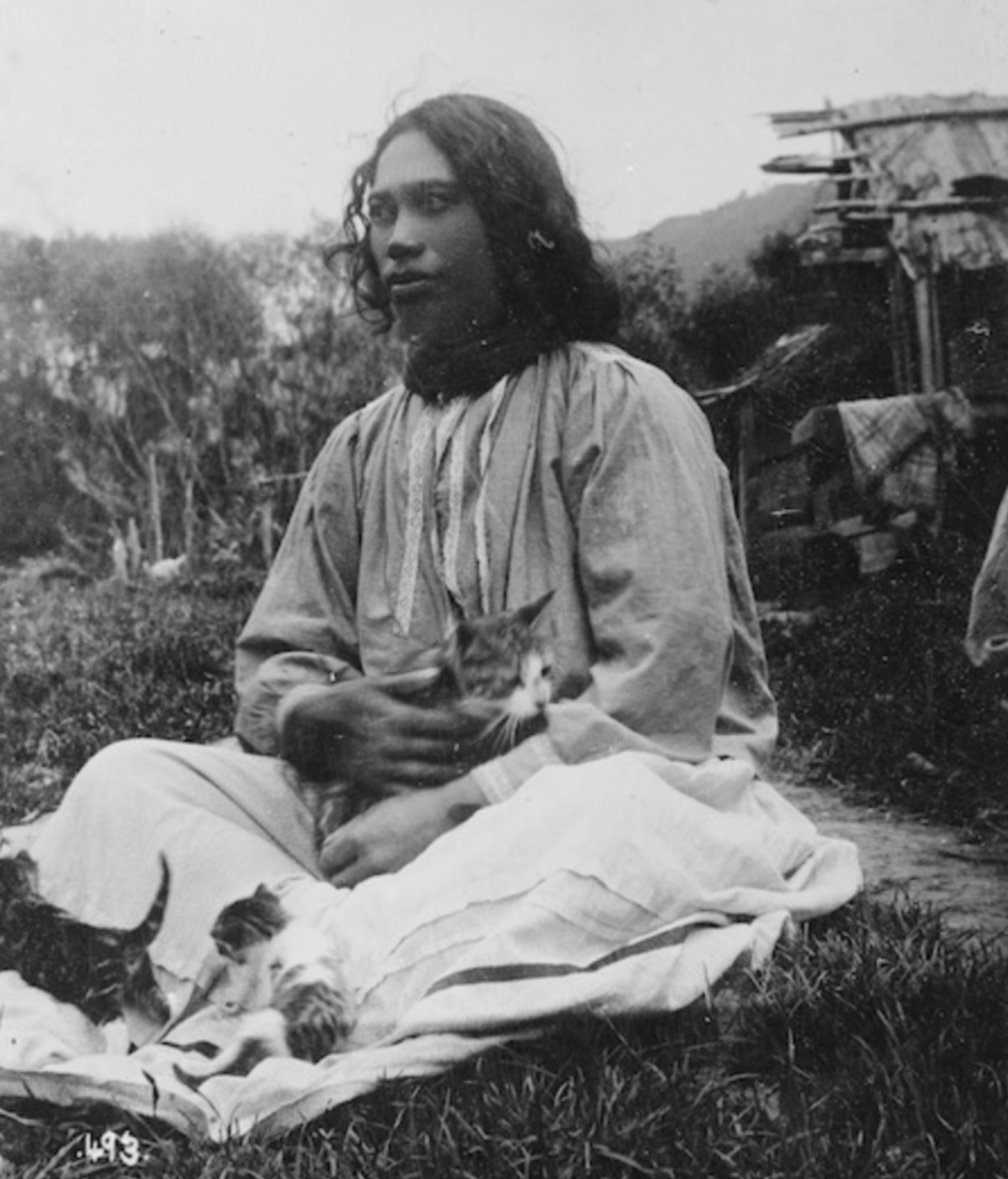 Black and white photo of a beautiful young woman with dark skin and black hair. She's wearing a loose blouse and skirt, sitting cross legged on grass. In her lap is a shorthaired tabby and white cat with a full tabby and a tabby and white kitten playing in the fabric of her skirt.