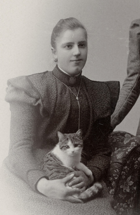 Black and white photo of a young white woman in Victorian dress, sitting in a chair fora studio portrait. Cradled in her lap is a white and tabby cat with the sweetest little face looking at the camera, its stripey tail wrapped around the woman's hands.