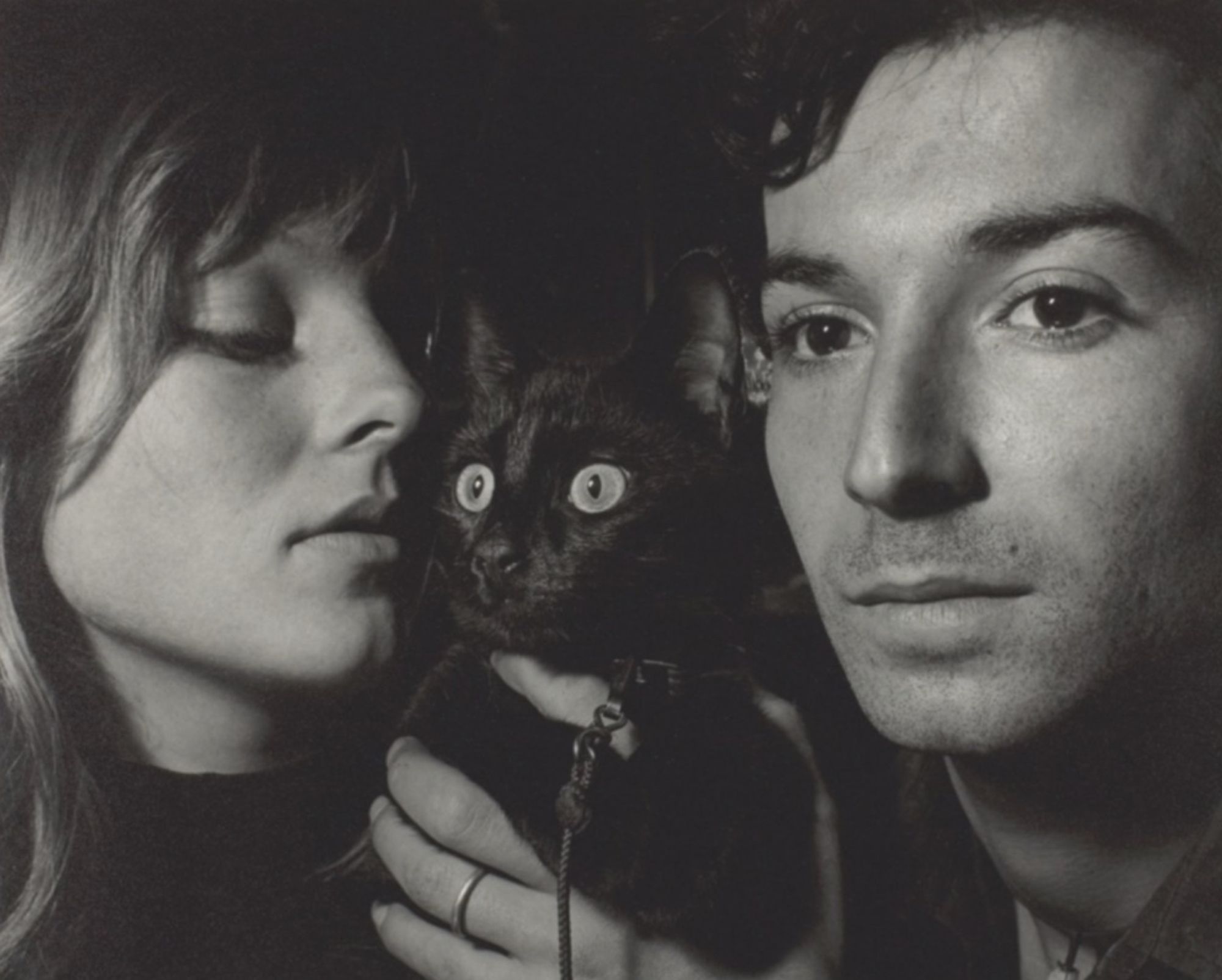 Black and white photo of an attractive young white couple with a shiny shorthaired black cat held up between them. It is a close up of all their faces and the cat looks very brightly into the distance as if it spotted something it wants to chase. The couple looks moody like they're in a French art film.