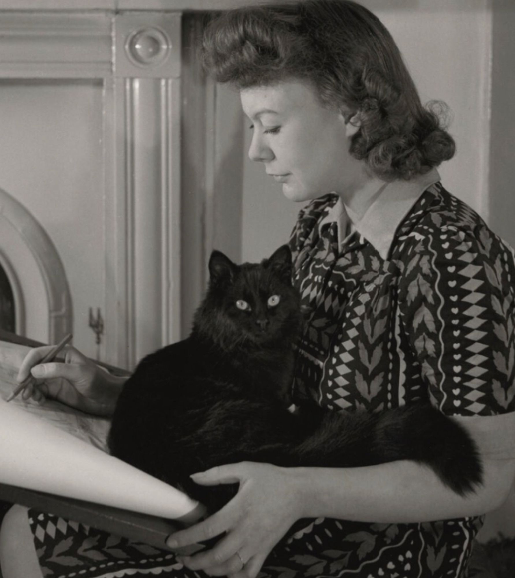 Black and white photo of a young white woman is badly patterned dress, working at a drawing table. In her lap is curled a gorgeous longhaired black cat that looks brightly up at the camera.