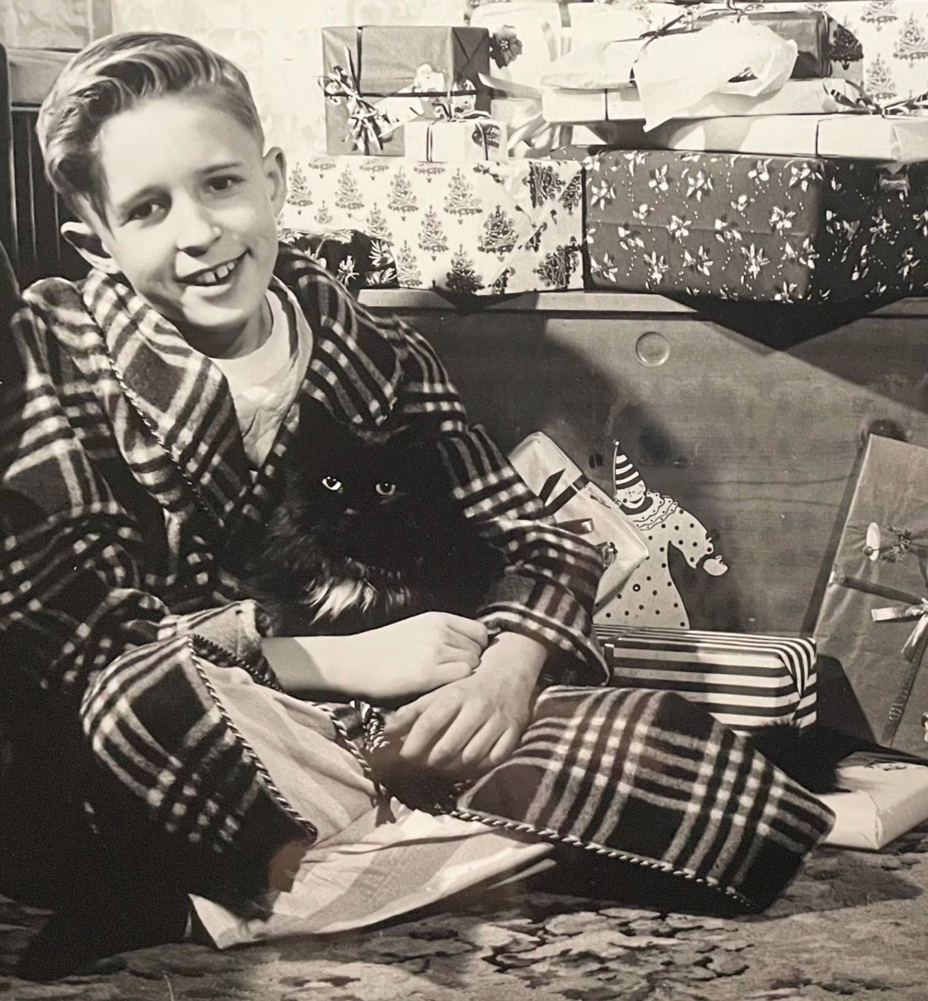 The same boy as previous photo, this time he is sitting in front of a large pile of Christmas presents, wearing a plaid bathrobe and pajamas, curled up in his lap is the same black cat!