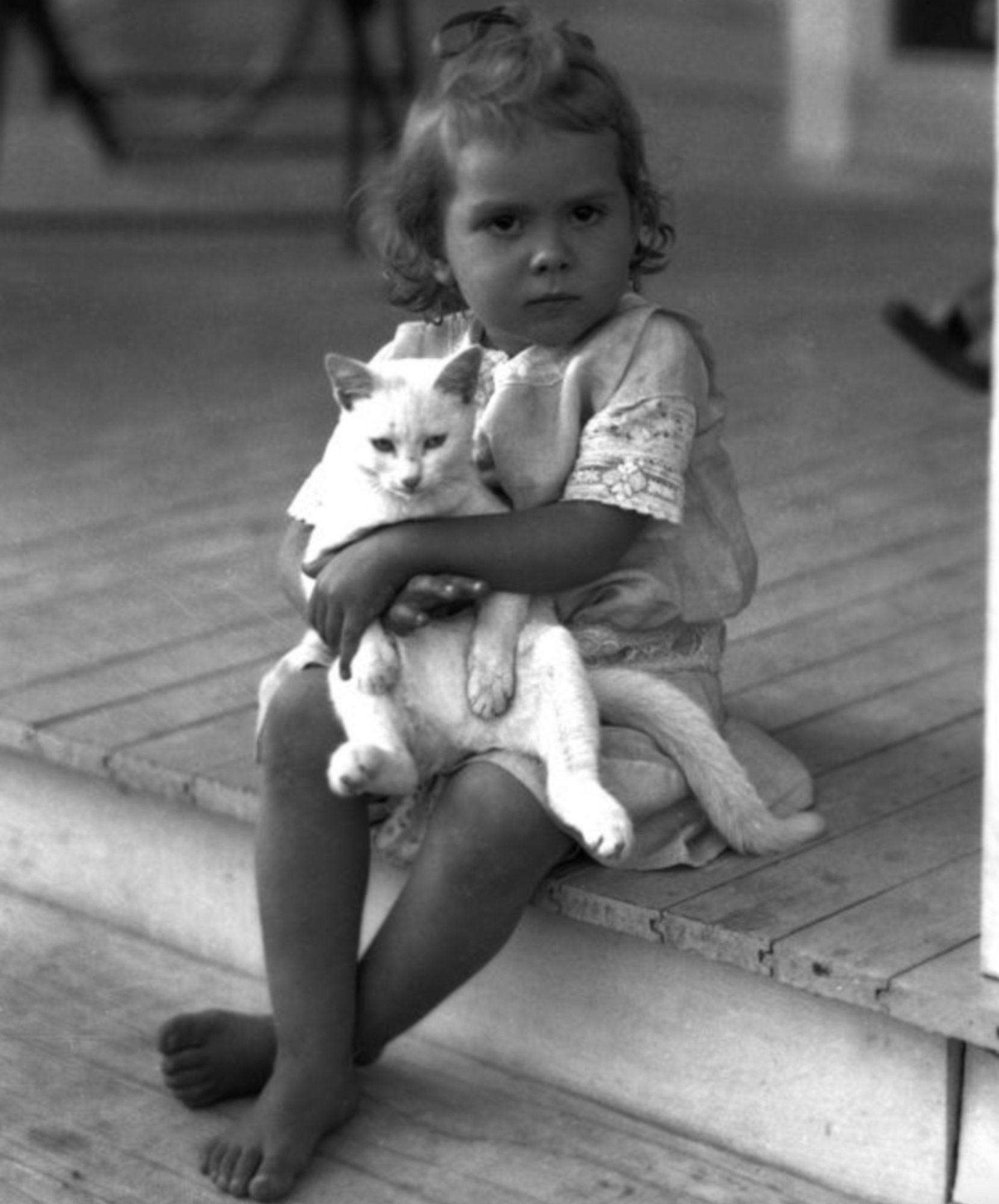 Black and white photo of a brown skinned little girl with loosely curly hair sitting on a wooden porch, holding a shorthaired white cat in her lap. The girl has a grumpy expression and the cat is sitting on its "butt", sort of upright in a human-like sitting position.