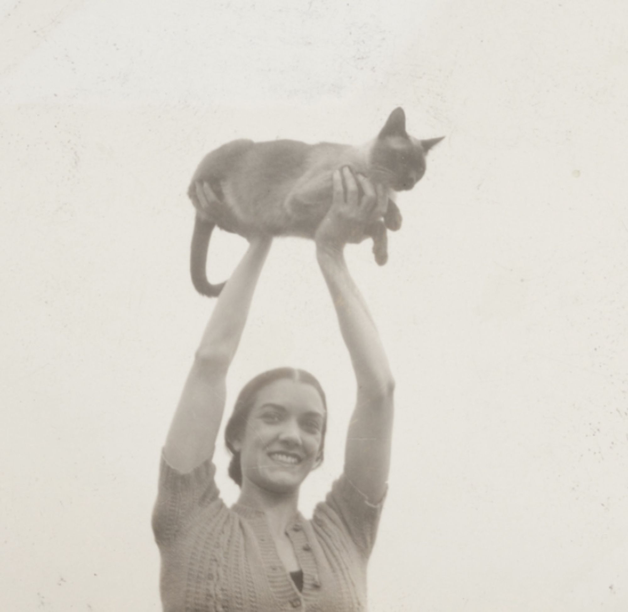 Black and white photo of a beautiful, slender white woman in a short sleeved cardigan, holding a Siamese cat aloft high in the air. The cat is sitting calmly in loaf position, firmly supported under its front and hind legs.