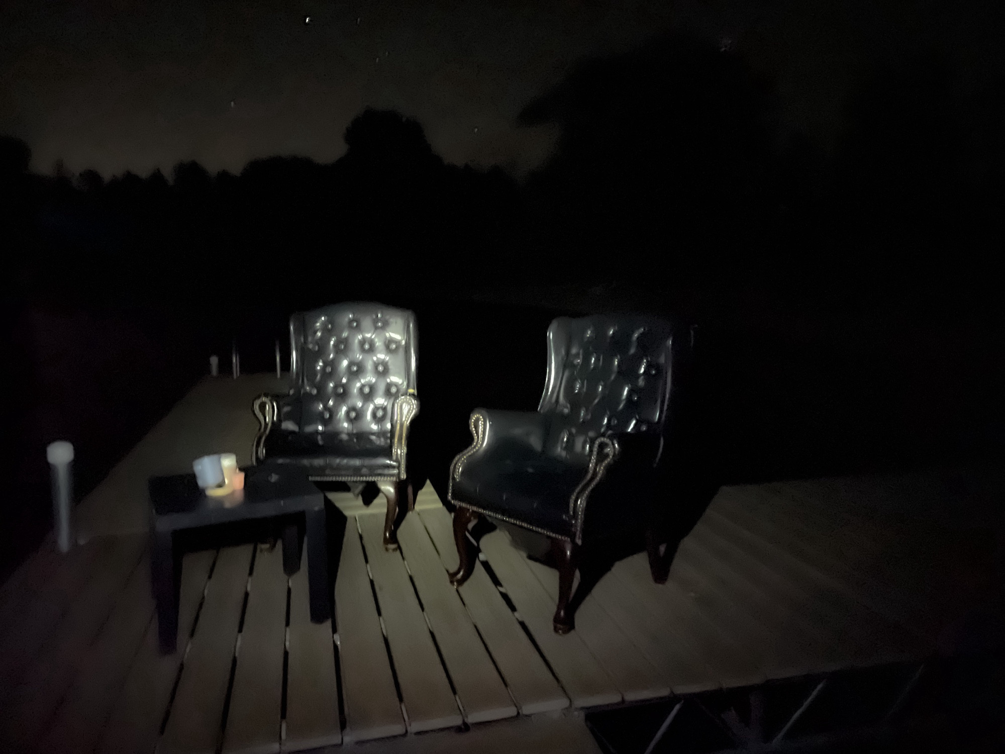 Creepy dark photo of some black fake leather armchairs sitting adjacent to a little table on a grey dock. You can see night sky behind them with a little light pollution glow.