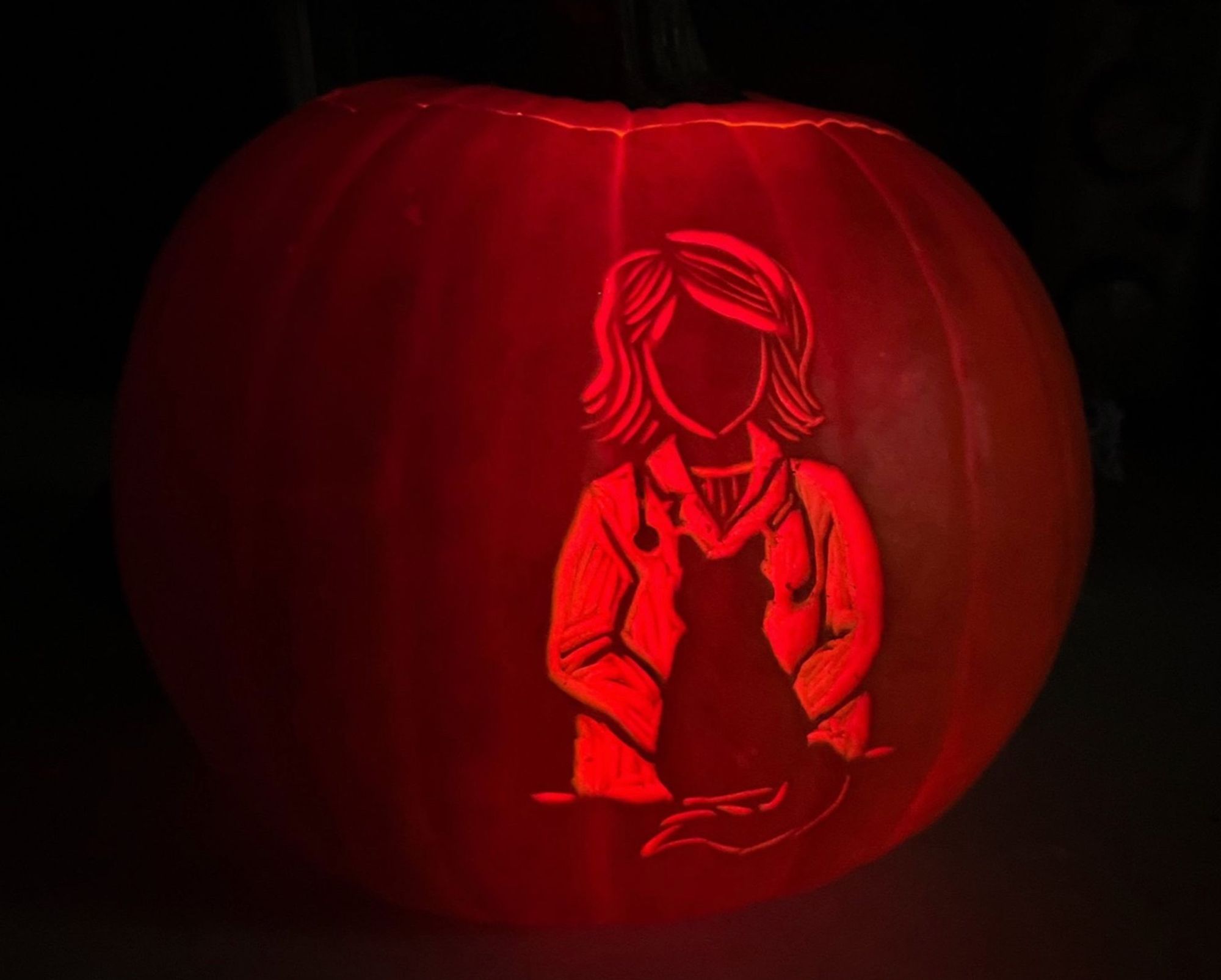 A small orange pumpkin in a very dark environment. The lighting from within it reveals a carving of a woman wearing a white doctor’s coat with a small stethoscope around her neck, in front of her sits the silhouette of a fluffy pear-shaped cat.