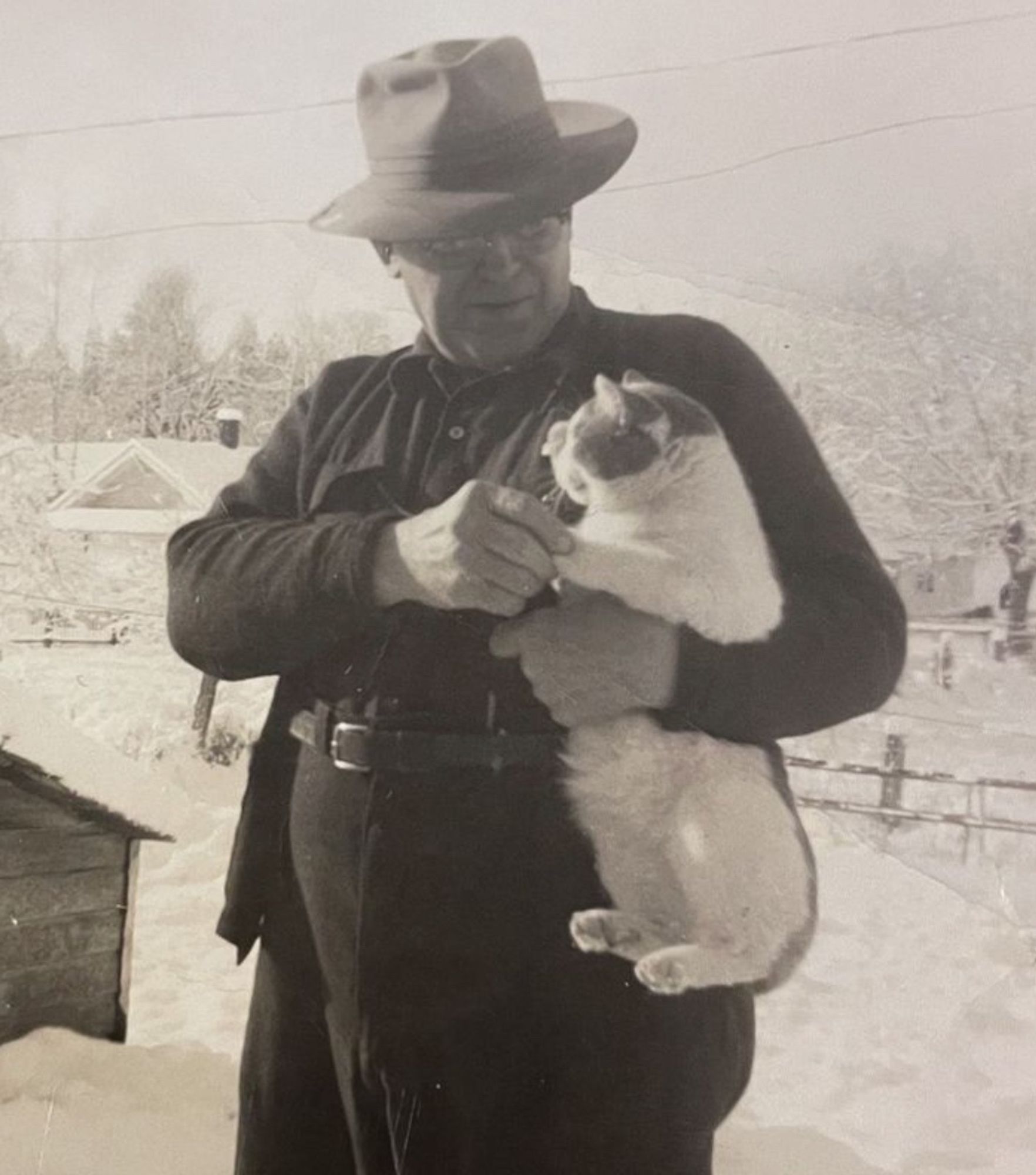 Black and white photo of an older white man in dark casual clothes and fedora type hat, standing outside in a beautiful snowy yard. He is looking affectionately down at a chubby shorthaired white cat with darker spots on its head that he cradles under his arm, its legs dangling down by his side. He’s gently holding one of the cats’ forepaws with his free hand and the cat seems relaxed and content.