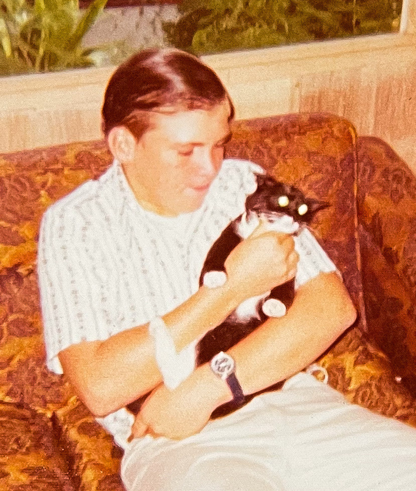 Color photo of a light skinned young man sitting on a brown and orange upholstered couch. His is cradling a black tuxedo kitten in his arms. The kitten appears to be trying to twist out of his arms and the reflection in its eyes is making them look like white laser beams.