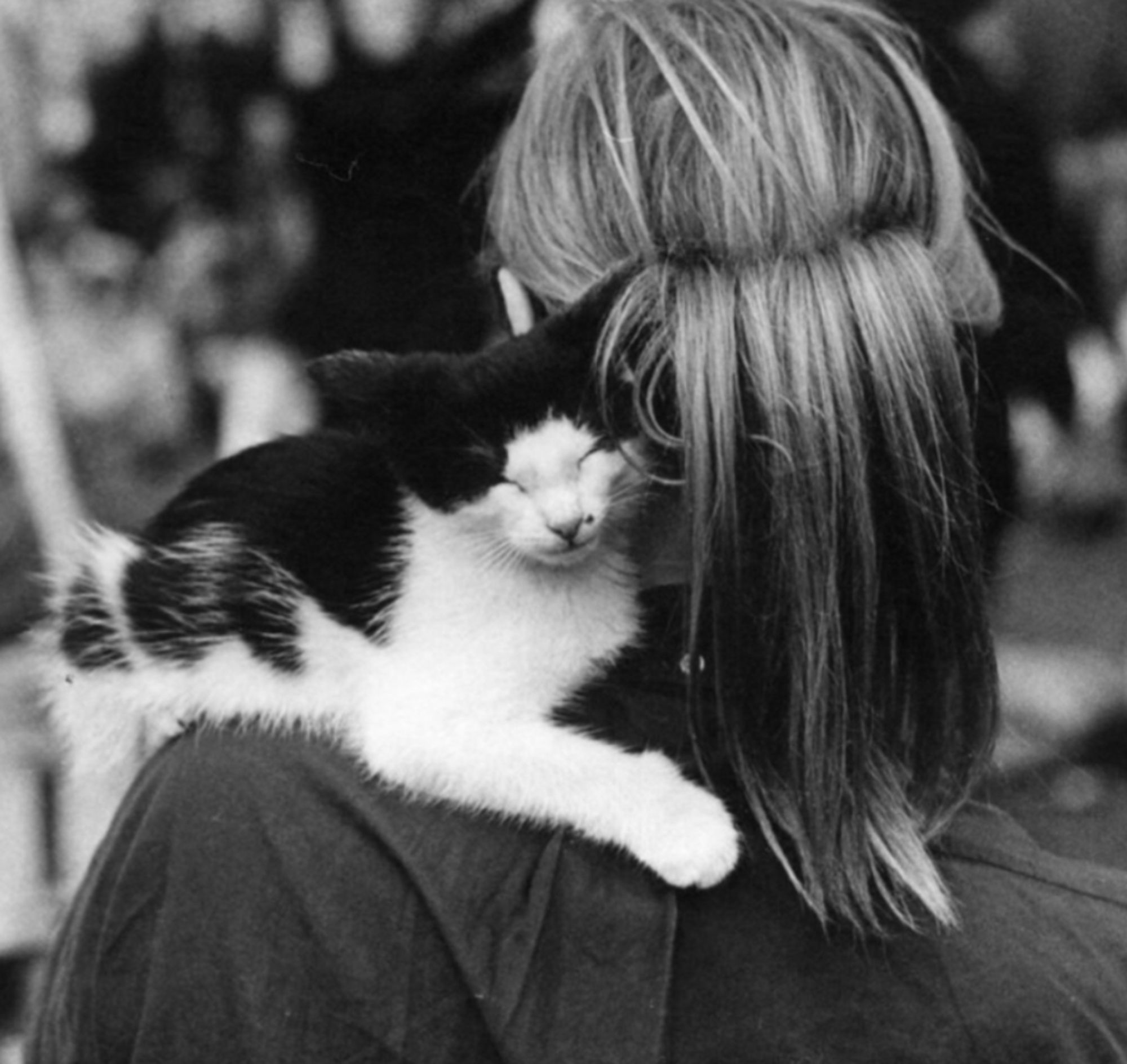 Black and white photo of an adorable black and white kitten that is sitting on a young woman's shoulder with its paw wrapped around the back of her neck, seeming fast asleep with its little face tucked against the woman's pinned up hair. The woman walking outdoors in a public garden, yet the kitten seems to be snoozing deeply.