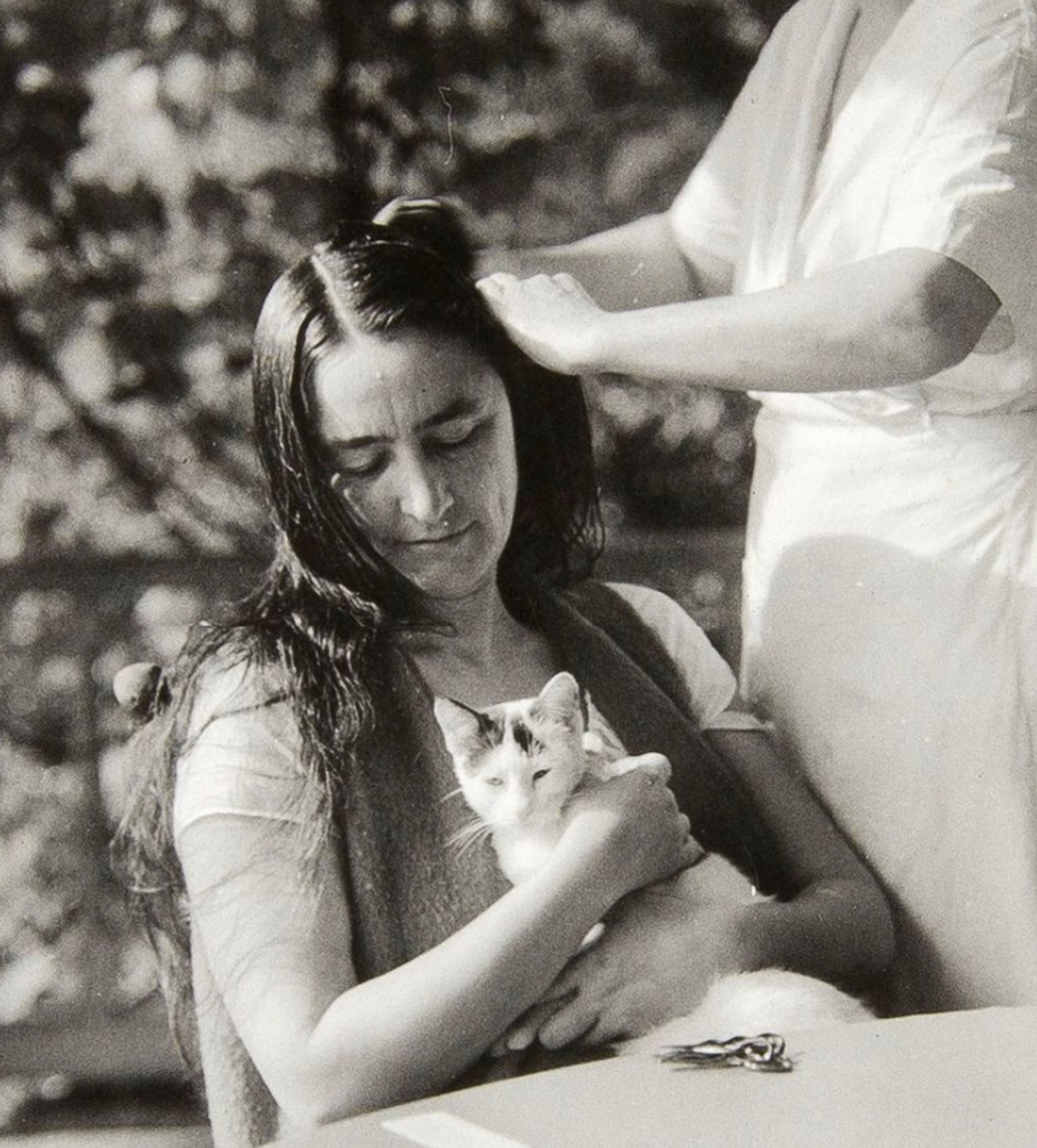 I'm going to copy and paste the description from the website because I think it's perfectly good: Portrait of Georgia O'Keeffe sitting in a chair at a table on the porch holding a white cat in her lap while getting her wet hair combed by a woman standing behind her who is wearing a white dress and apron (possibly Margaret Prosser); O'Keeffe sits with her head turned to the left looking fondly down at the cat, she is dressed in a white short sleeved scoop-neck blouse with a fuzzy-textured vest; blurred foliage and porch railing in the background