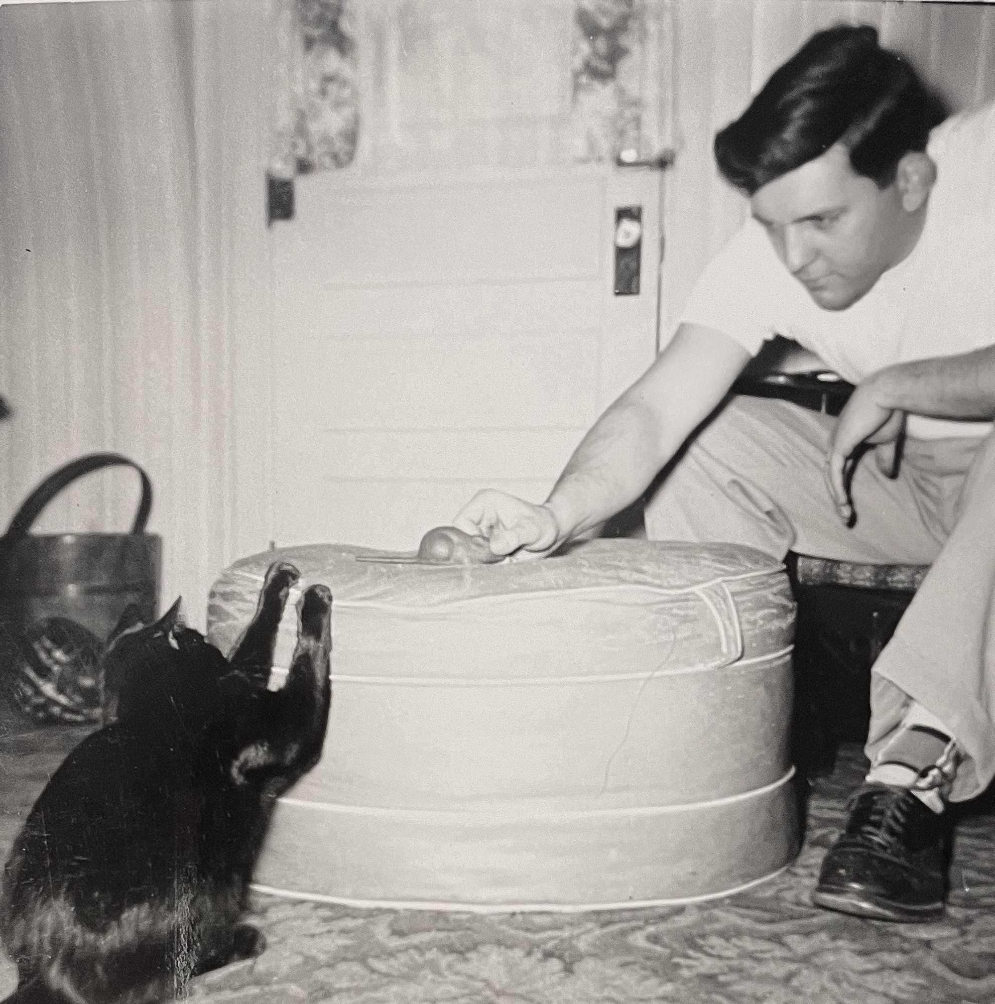 Black and white photo of a young white man with dark hair, leaning forward from his seat to move a mouse toy on an ottoman in front of him. In the opposite side of the ottoman is a shiny, shorthaired black cat that has both forepaws excitedly braced on the edge of the ottoman, ducking down slightly as if hiding from the mouse.