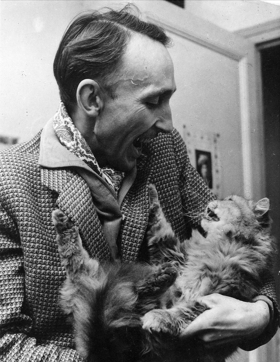 Black and white photo of a thin white man wearing a paisley ascot, corduroy shirt, and sports coat made of a patterned tweed of some kind? I don't know what it is. It created the effect of a tiny houndstooth-ish pattern. The man is joyfully looking down at a longhaired tabby in his arms, which he is holding upside down like a baby. Both the man and cat have mirrored, open-mouthed expressions.