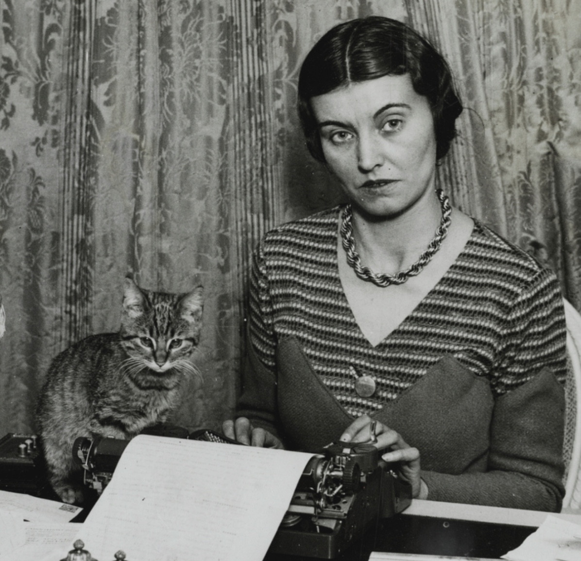 Black and white photo of a nicely dressed woman sitting at a paper-strewn desk, seeming to type on a typewriter. Beside her sits a shorthaired tabby cat and both of them look quite severely toward the viewer.