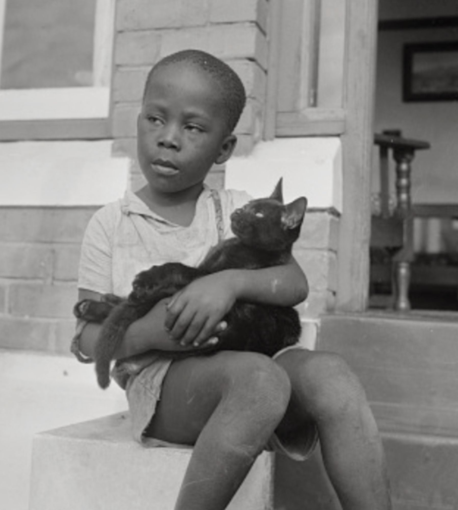 A young black boy sits on the stoop of a house with a very serious expression on his face. Cradled in his arms like a baby is a very content-looking shorthaired black cat.