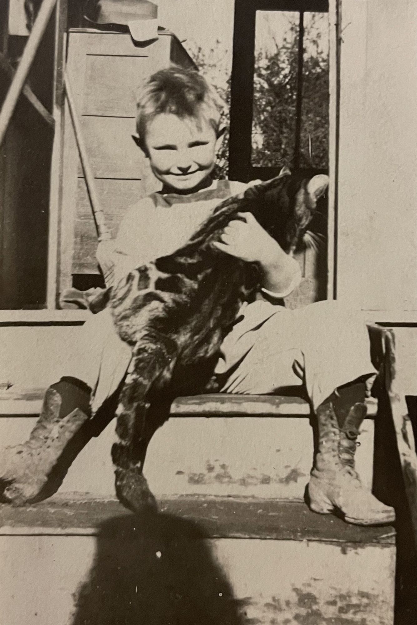 Black and white photo of a young white boy sitting on the steps of a wooden house, smiling at the camera and awkwardly holding a large shorthaired tabby cat.