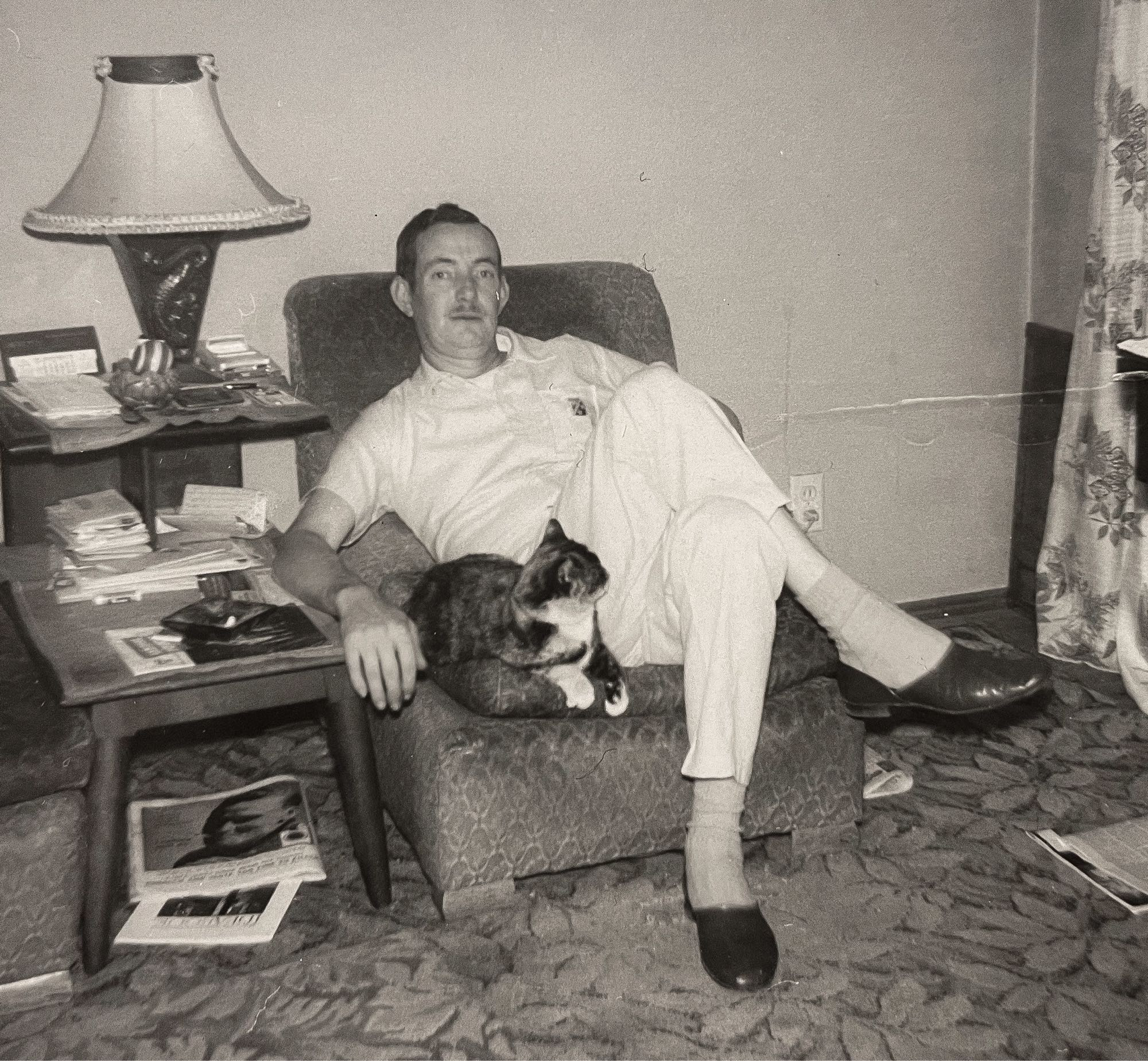 Black and white photo of a white middle aged man sitting in an upholstered armchair in a living room type setting. A cute little shorthaired calico cat is squeezed onto the chair cushion beside the man, both of them looking comfortable sharing the chair together.