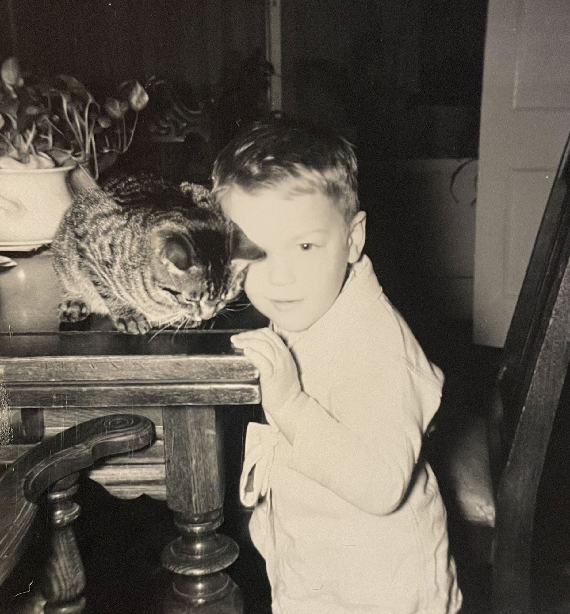 Black and white photo of the same cat and boy from the previous photograph. This time the cat is sitting on the edge of a table and the boy is standing next to it, his head leaning affectionately into the cat's side.