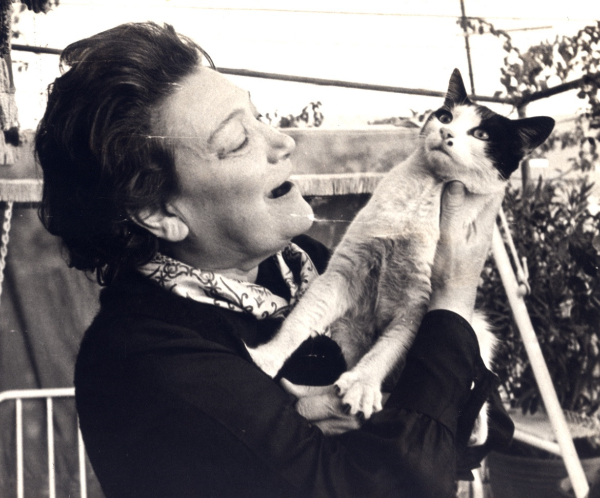 Black and white photo of a 62 year old Greek woman standing in a backyard, holding a shorthaired white cat with black patches. The cat is pushing away from her with a look of disdain.