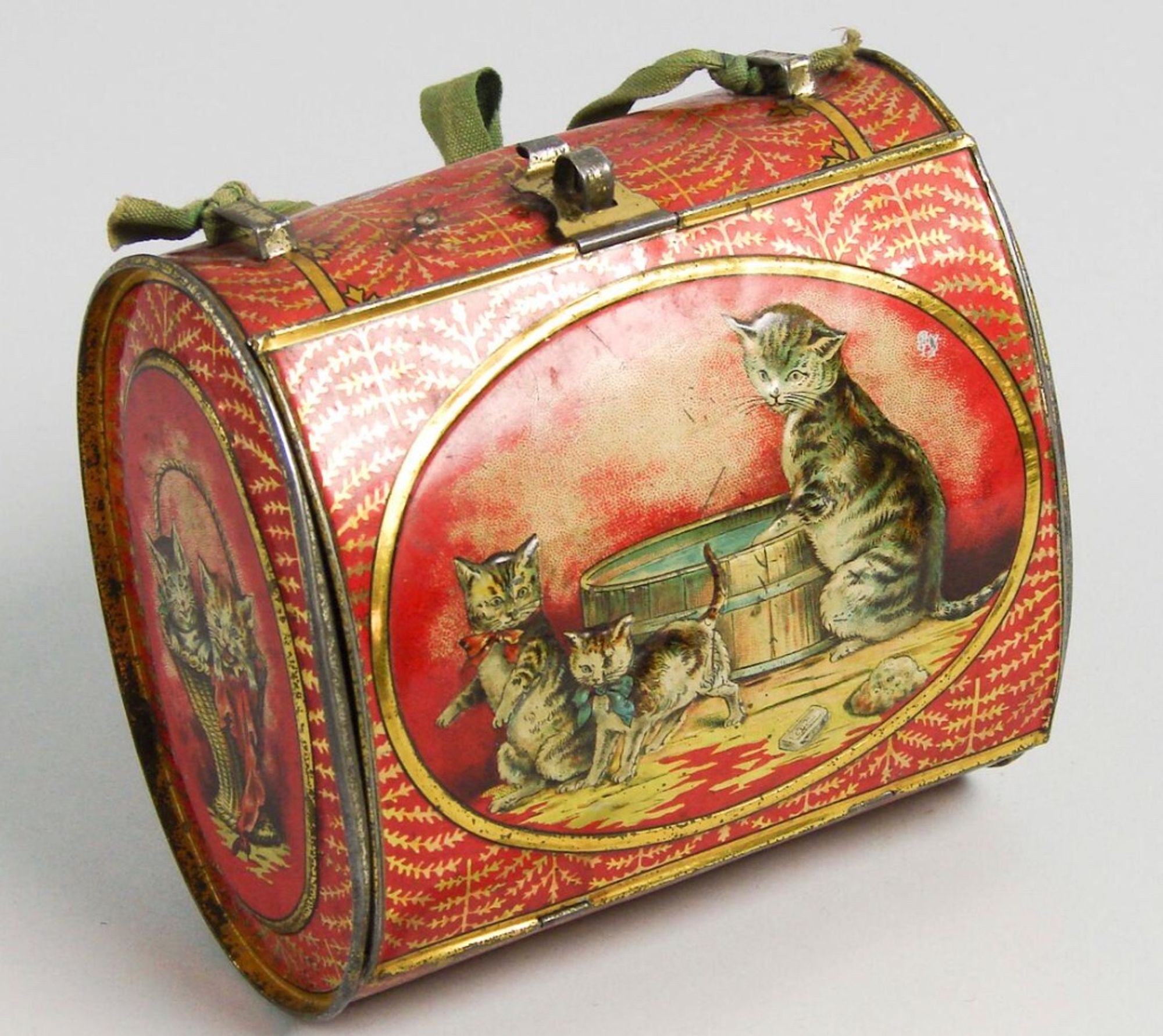 Color photo of an oval shaped metal carrying case, like a little tin purse for children. It's painted red with delicate gold details all over and a large illustration on the front. The illustration depicts a shorthaired tabby mother cat that seems to be washing something at a basin of water while looking over her shoulder at her two kittens. The kittens are running off, a tabby one wearing a red bow and a tabby and white one wearing a blue bow.