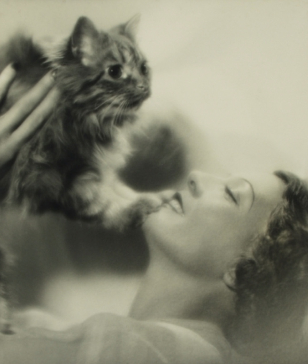 Dreamy, slightly hazy black and white profile photo of a white woman in flawless makeup, smiling ecstatically while lifting a longhaired tabby cat up above her face. The cat has braced its paws on the woman's chin and looks brightly off into the distance.