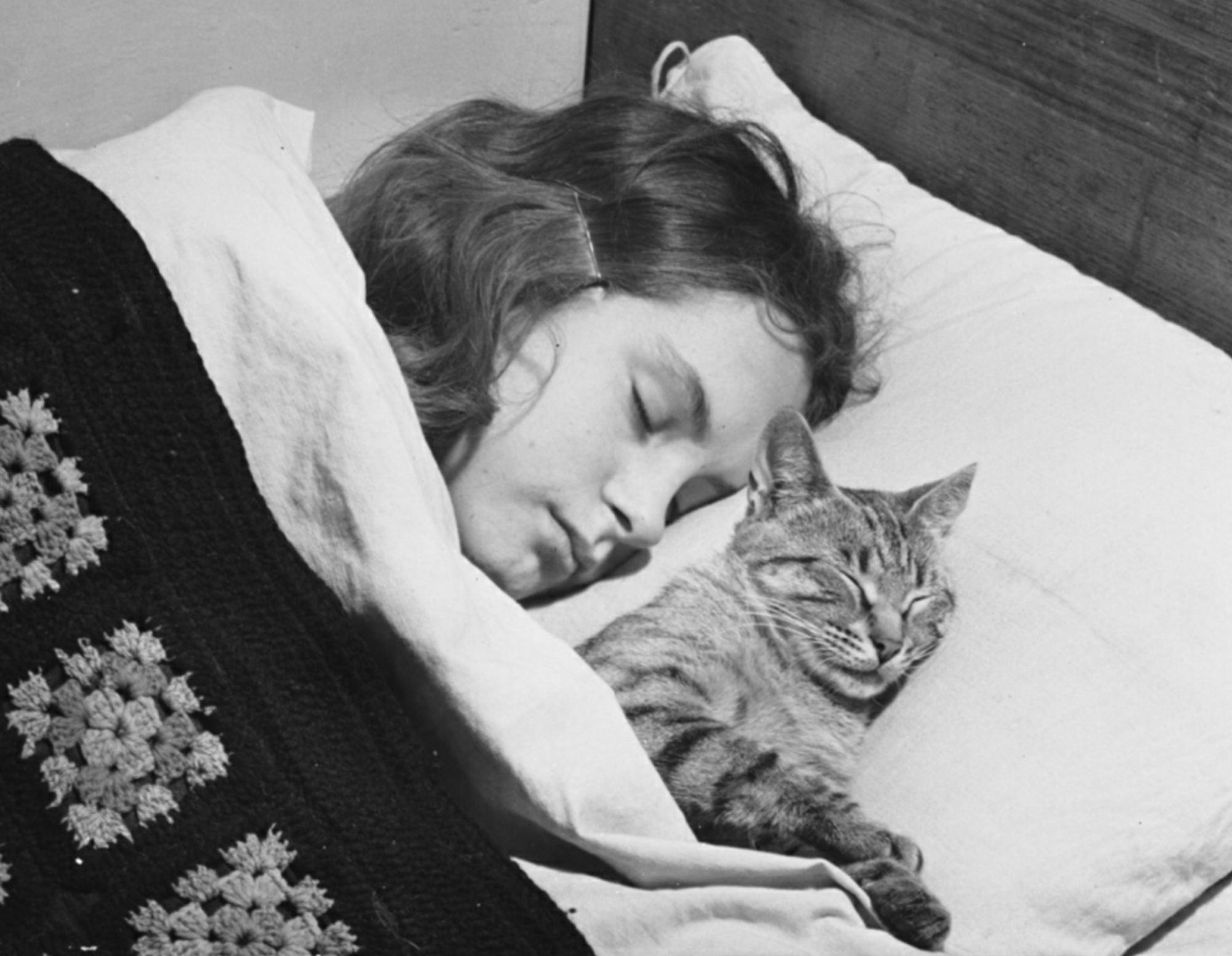 Black and white photo of a young white girl lying in bed with her eyes closed, tucked under a sheet and crochet square blanket. Next to her, sharing her pillow and also tucked under the covers, is a sleeping shorthaired tabby cat.