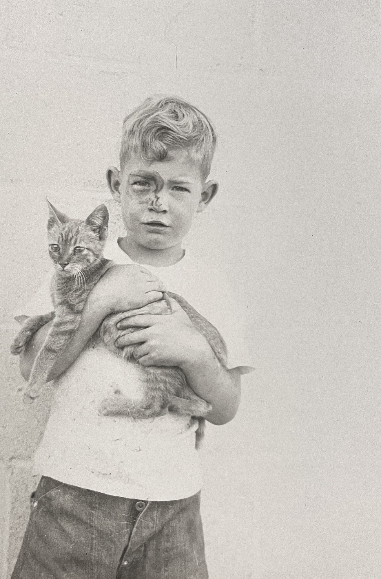 Black and white photo of a blonde white boy in white tee shirt and darker pants, cradling a shorthaired tabby kitten in his arms. The boy looks at the camera and has some kind of face paint around his right eye.