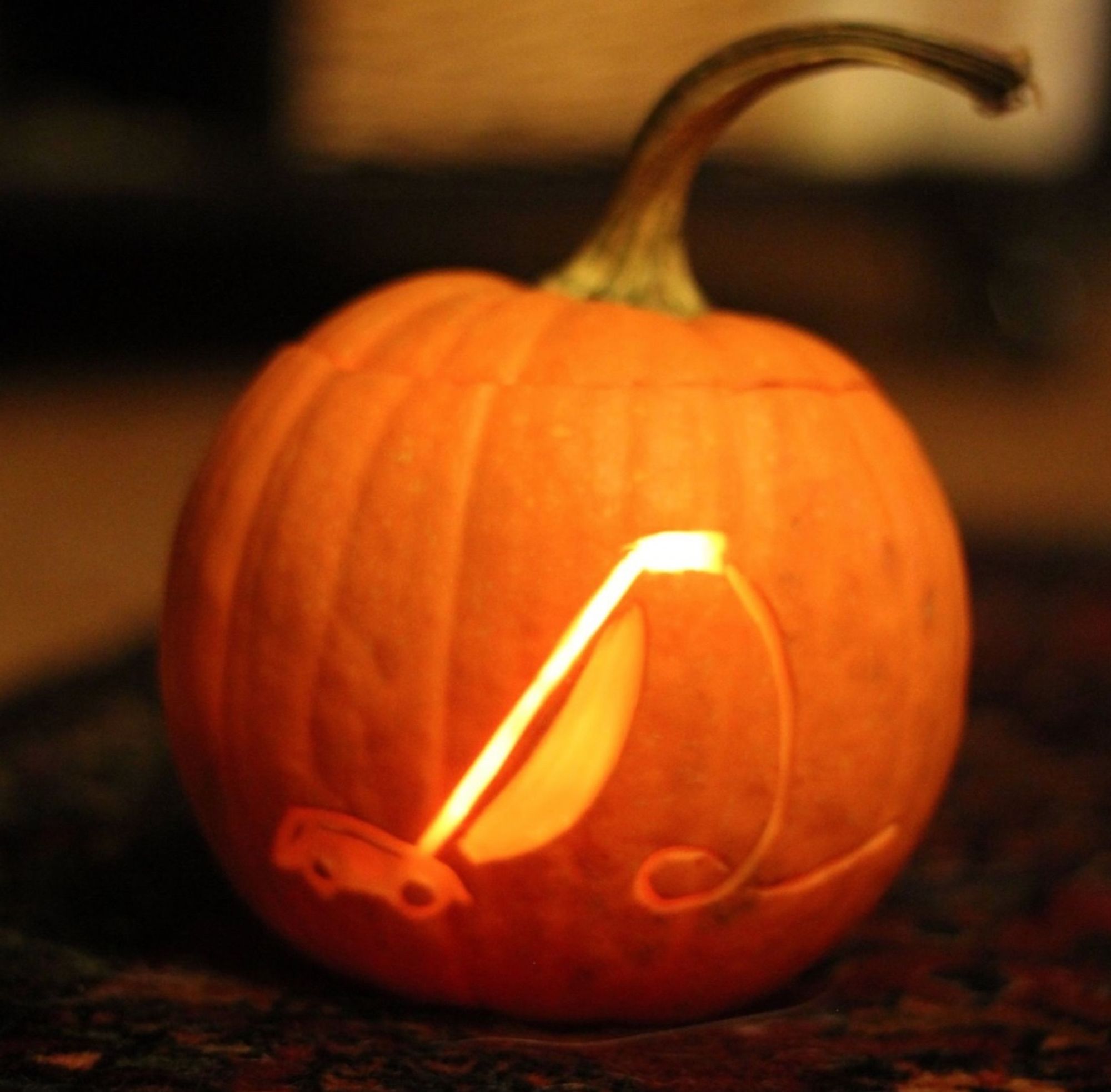 An orange mini pumpkin with an upright vacuum carved into with its coiling cord trailing away. It's somewhat dark and the pumpkin is illuminated from within.