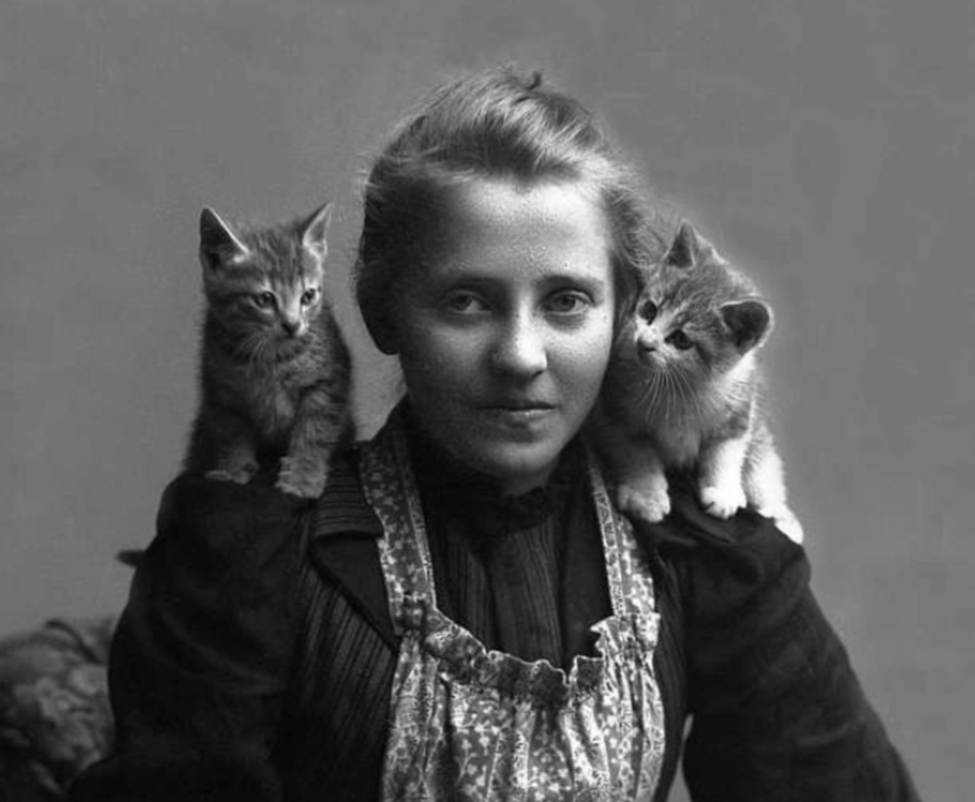 Black and white studio portrait of young white woman in a dark dress with floral print apron. On each of her shoulders sits a shorthaired kitten, one tabby and one tuxedo tabby.