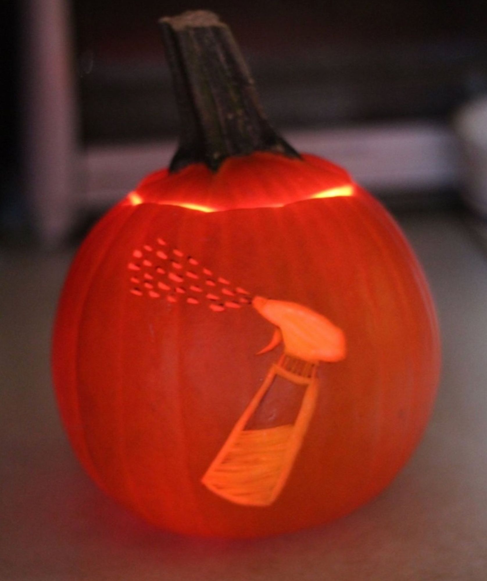 An orange mini pumpkin with a spray bottle carved into it, dashed lines represent water coming out. It's somewhat dark and the pumpkin is illuminated from within.