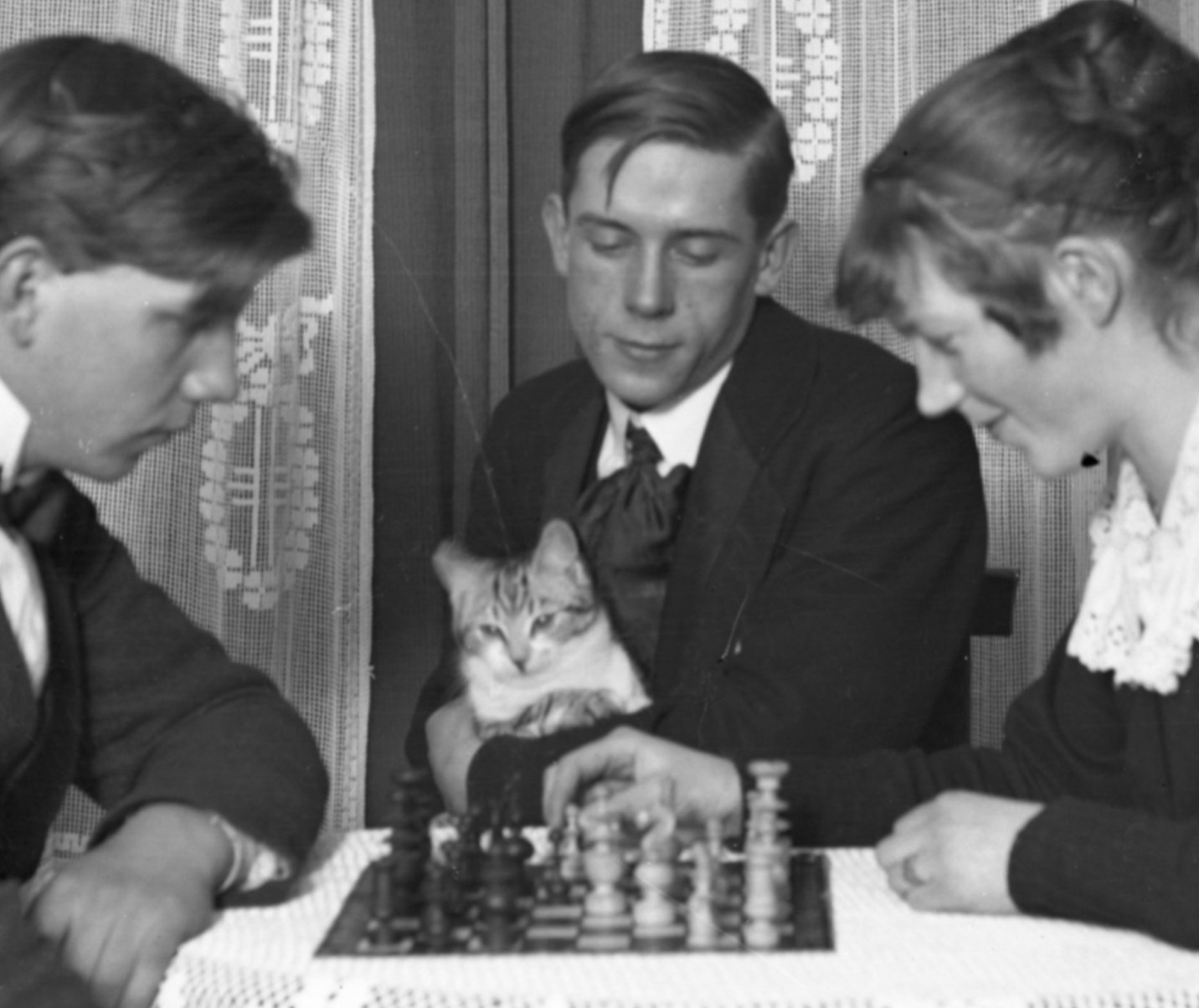 Black and white photo of a white young man and white young woman playing chess across a table with white lace table cloth. Facing the camera is another white young man who is watching the game with a shorthaired tuxedo tabby kitten in his lap. The kitten also appears to be watching the game.