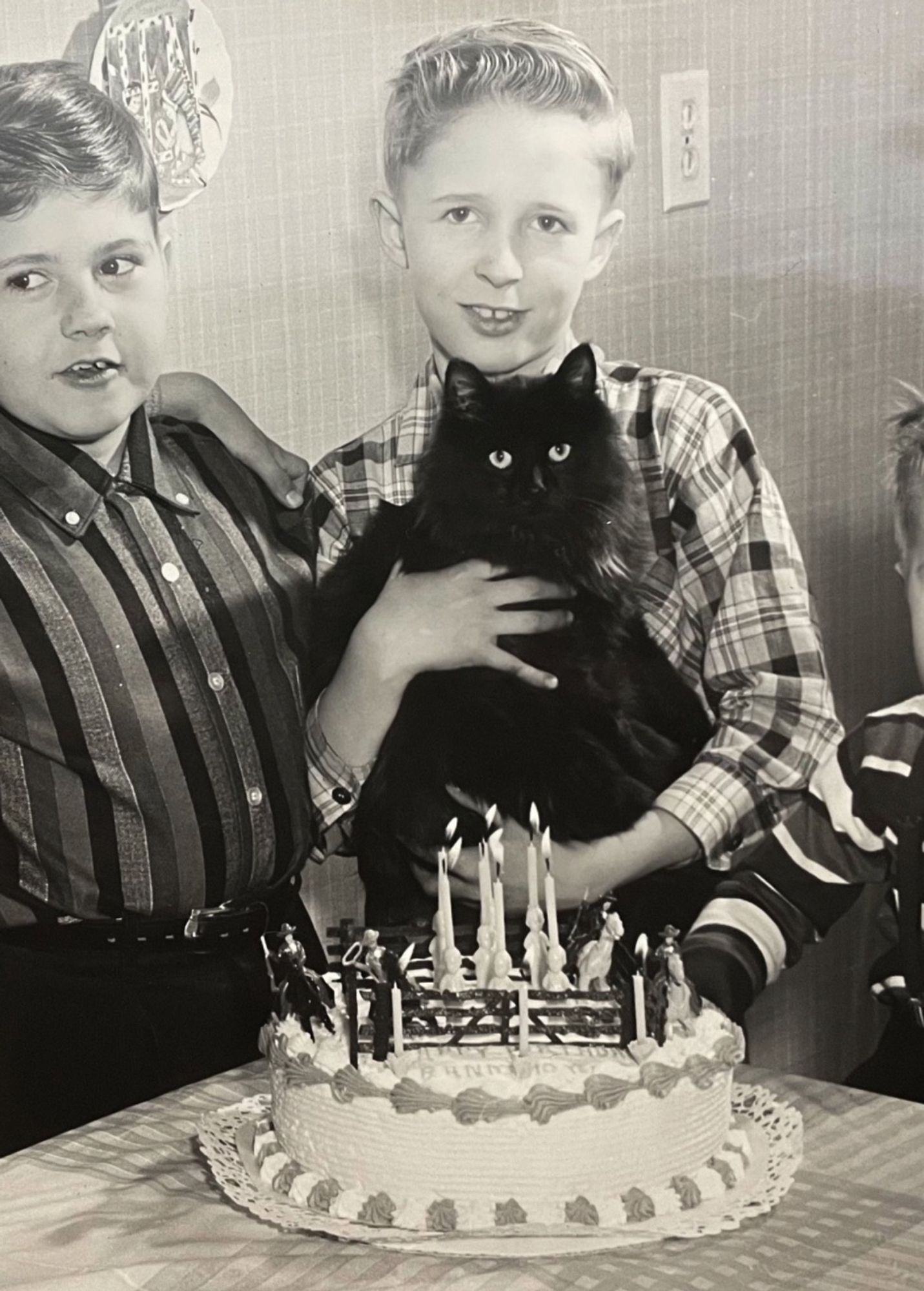 Black and white photo of a smiling blonde, white boy holding a beautiful longhaired black cat. He’s standing at a table with a cowboy-themed birthday cake in front of him and other boys on either side.