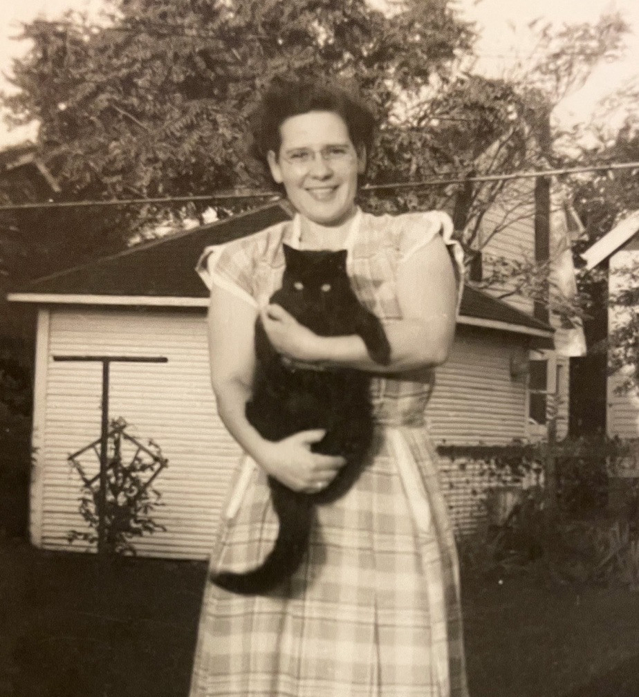 Black and white photo of a young white woman in a plaid summer dress, standing outside in what looks like a backyard. She smiles at the camera and holds a hefty shorthaired black cat in her arms.