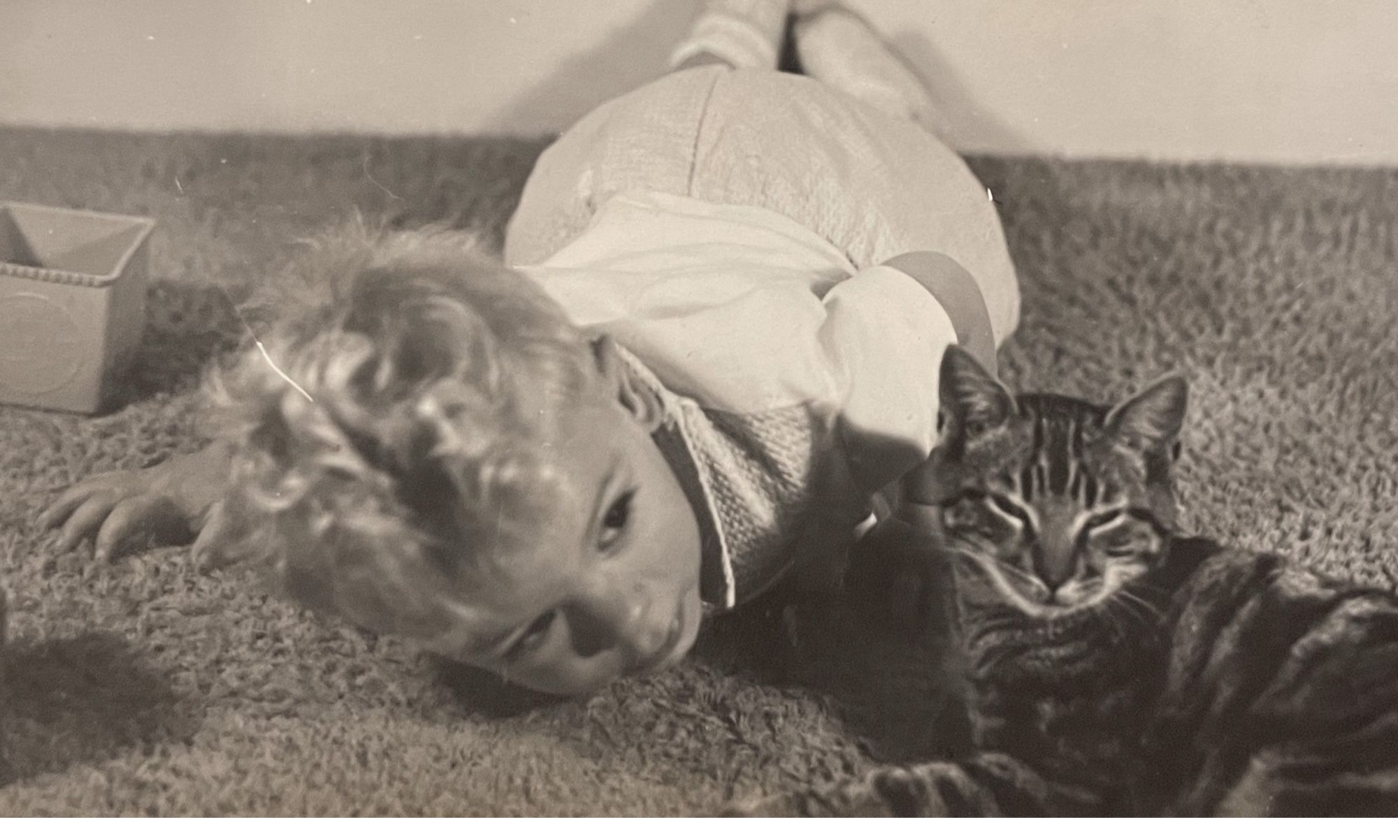 Black and white photo of a blonde, white toddler laying on a carpet with its head next to a shorthaired tabby cat that is sitting relaxed nearby.