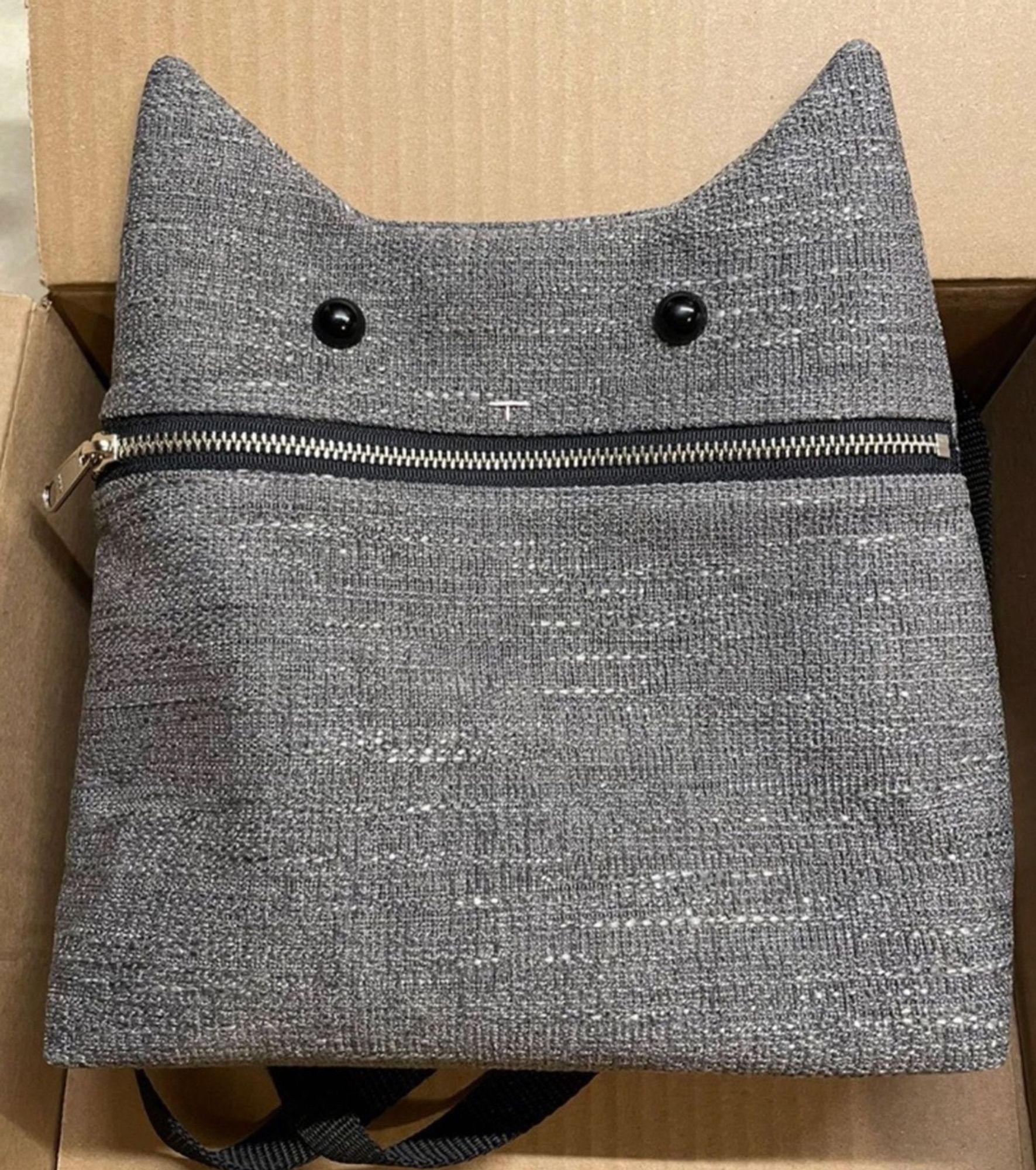 Photo of a grey purse that is in the square, stylized shape of a cat face. Two shiny black buttons are the eyes, a zipper forms the mouth, and pink stitches make a minimalist pink nose.