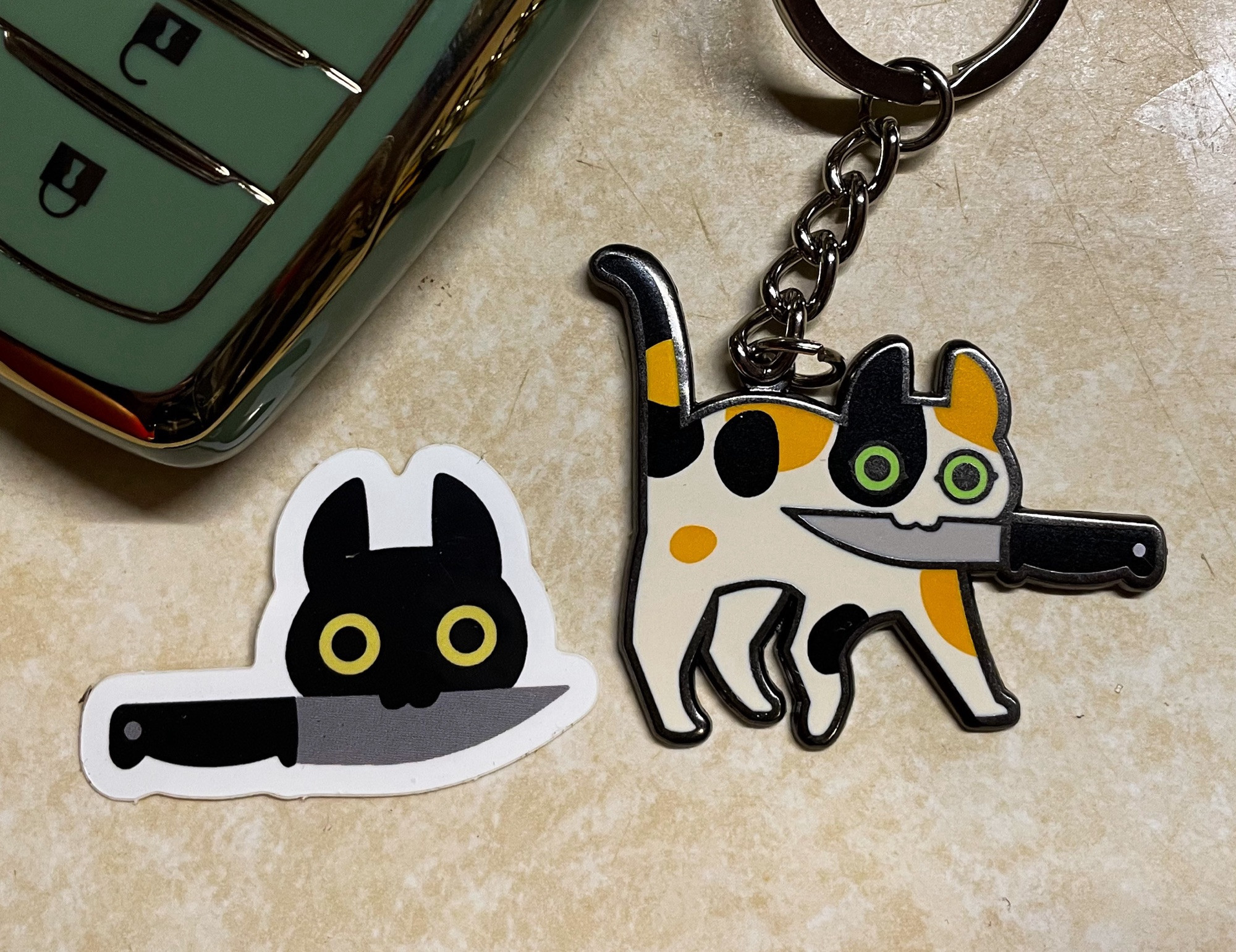 An enamel keychain depicting a cute green eyed calico cat holding a knife in its mouth. Next to it is a little sticker showing a black cat’s face holding a knife in its mouth.