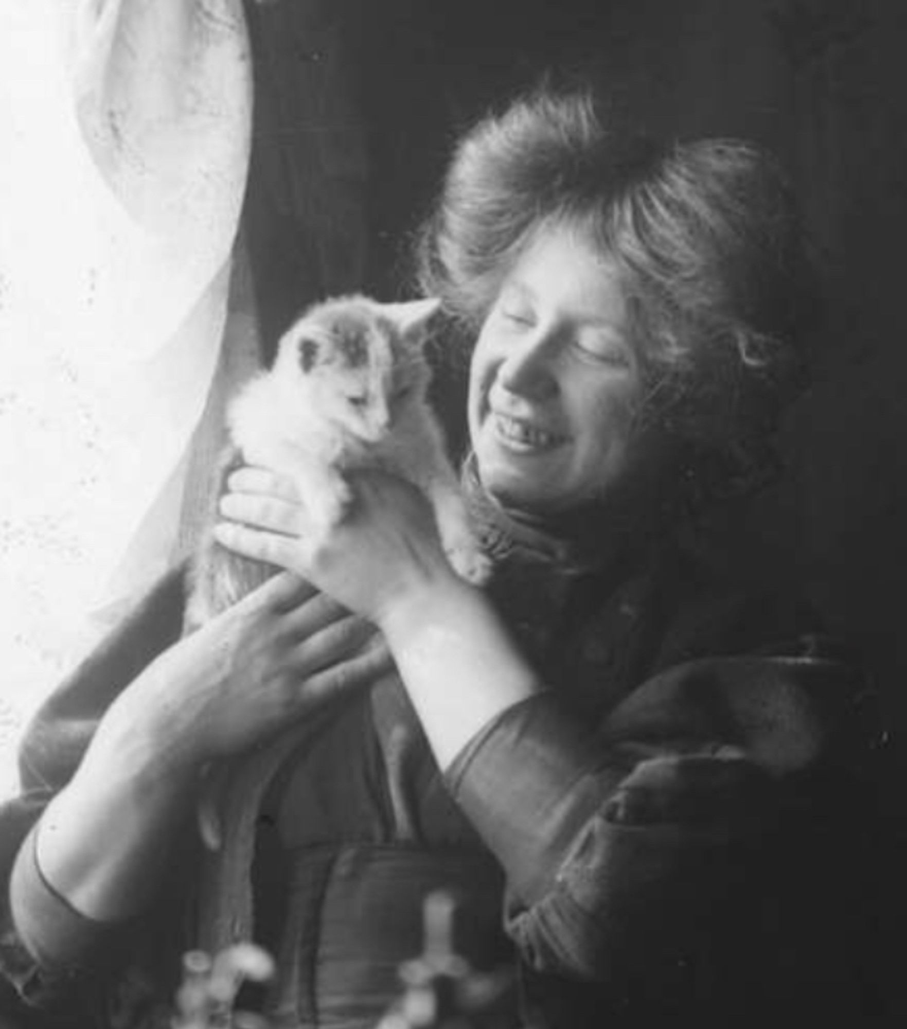 Black and white photo of a young white woman with her haired styled into two symmetrical mounds on her head. She is seated by a sunny window, illuminated by the cheerful light coming through the brilliant white curtain, it lights up her face and the grumpy looking white and spotted shorthaired kitten she hold up in her hands.