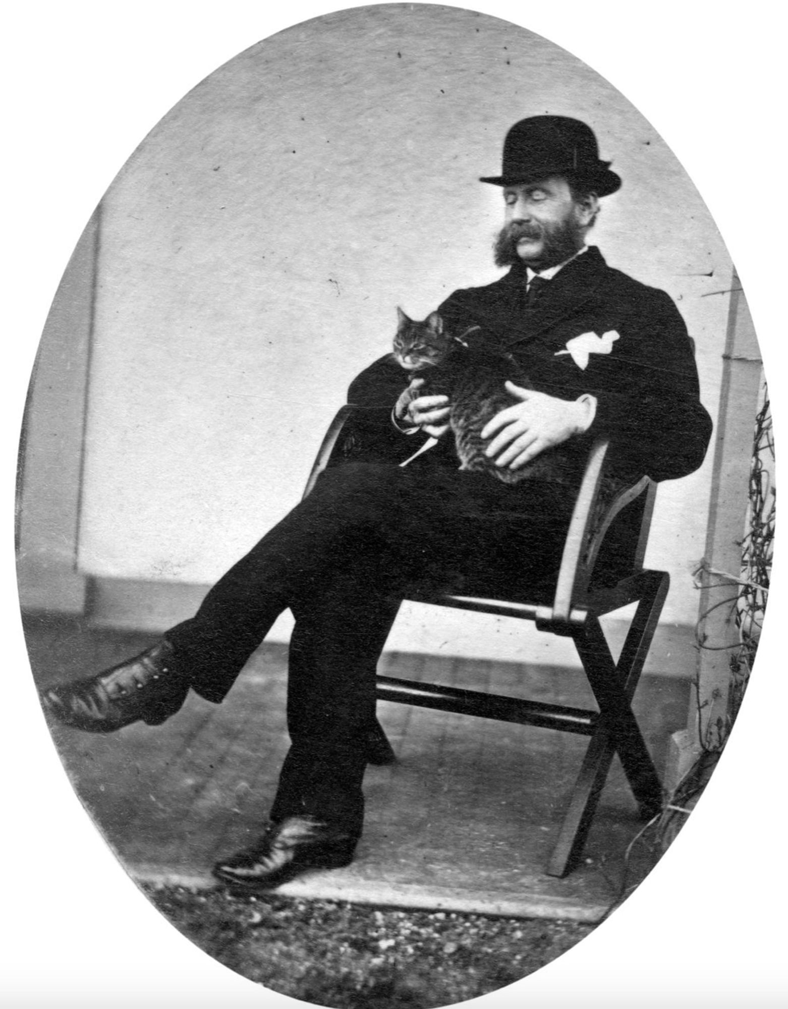 Black and white photo of a dapper middle aged man with thick muttonchops facial hair in a beautiful dark suit, bowler hat, and finely crafted leather shoes with buttoned gaiters. He is sitting in a folding wooden chair outside on a porch, smiling down at a shorthaired tabby cat he is gently holding in his lap.