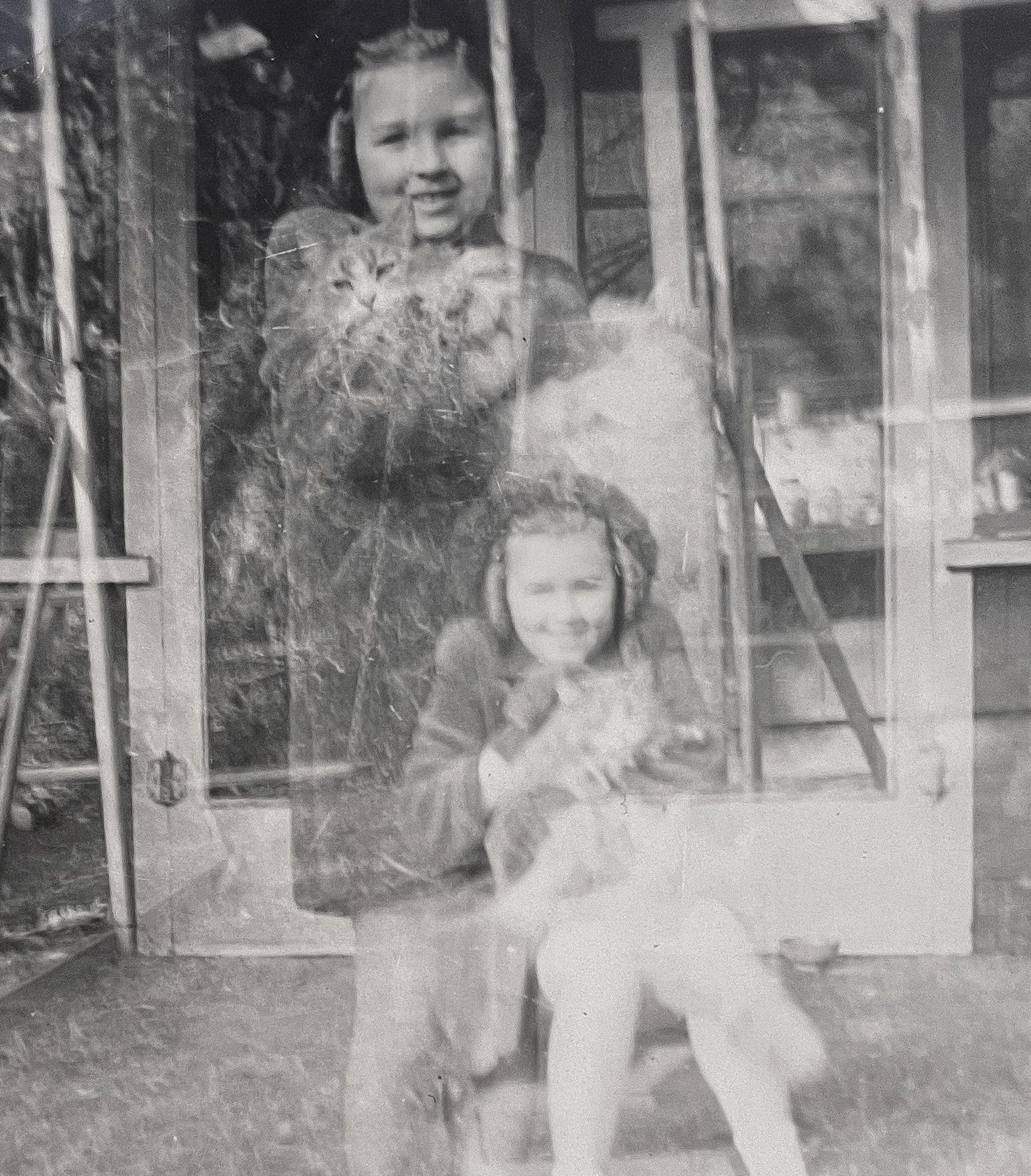 Two white girls in cold weather outfits, posing for a photo, each holding a longhaired cat. They appear to be somewhat translucent apparitions on a wooden house porch due to a double exposure on the film.