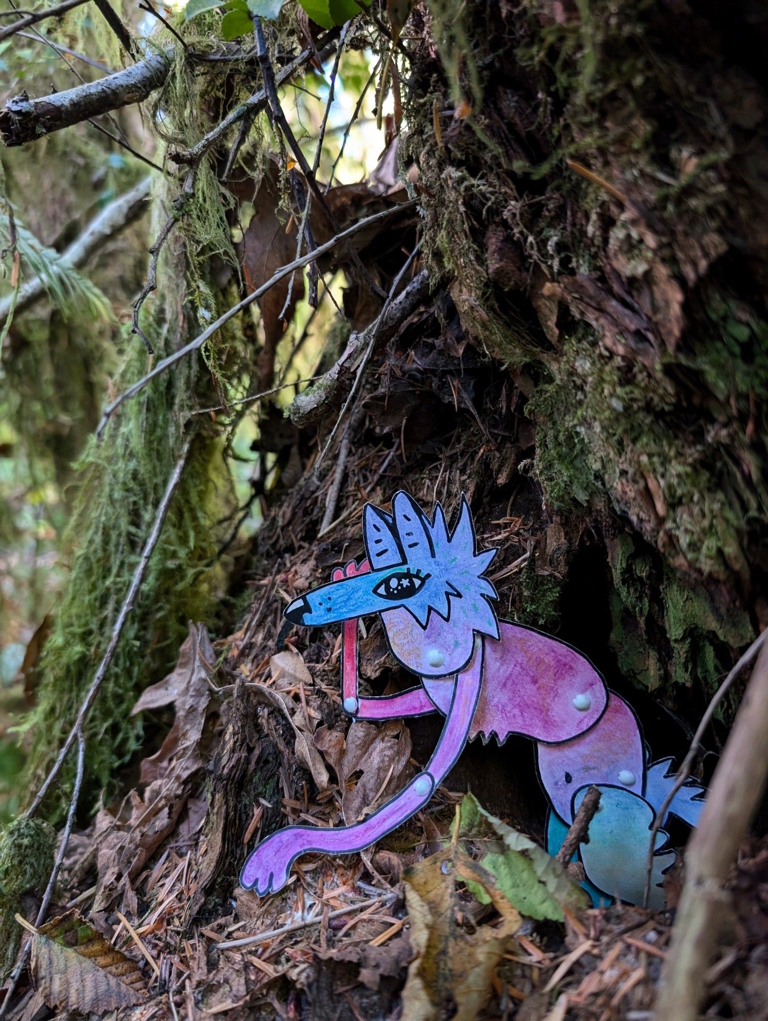 A colorful paper coyote character peeks out of a hole in a stump among the leaves and twigs