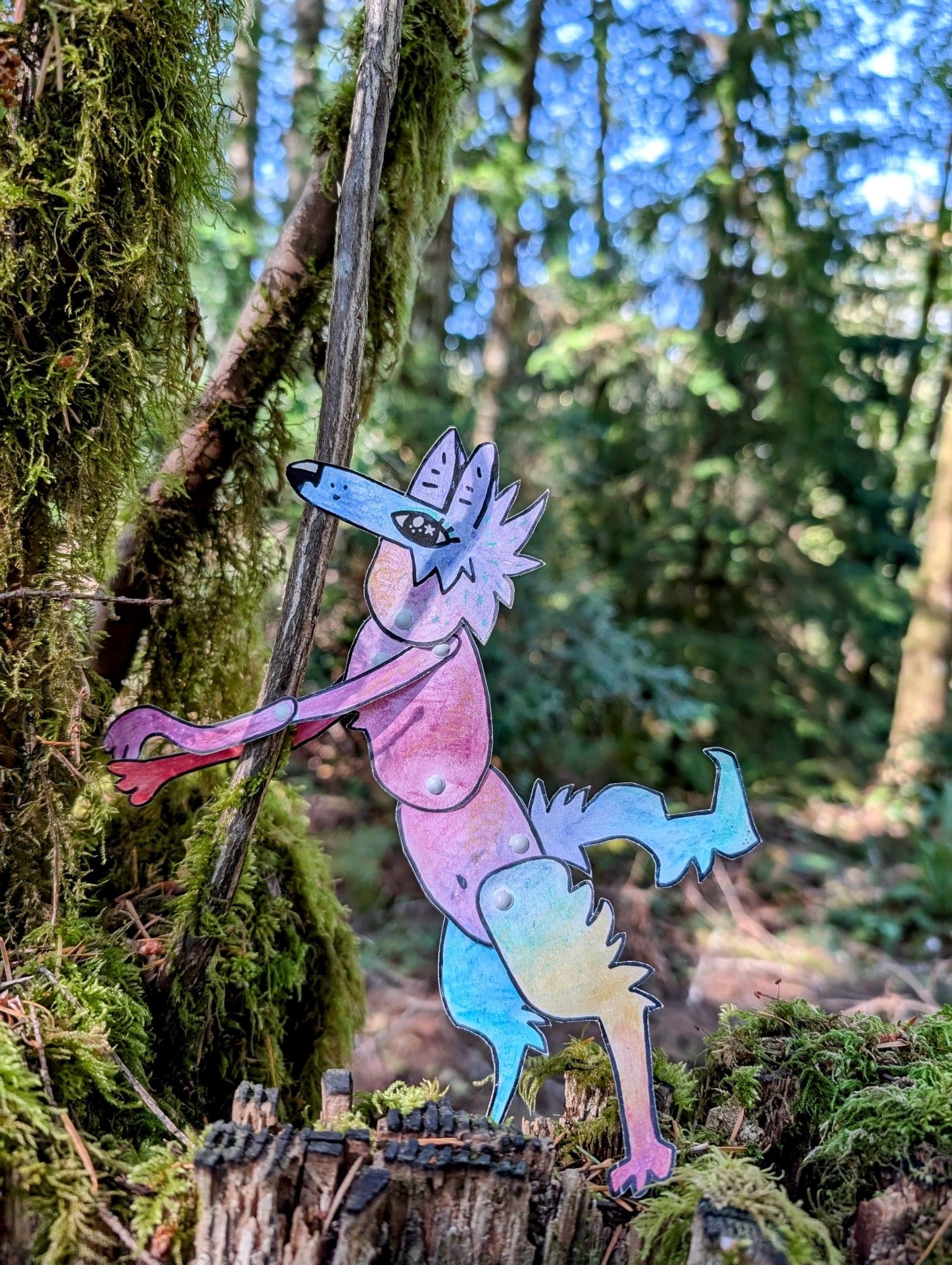 A colorful paper coyote character leaning against a small mossy branch