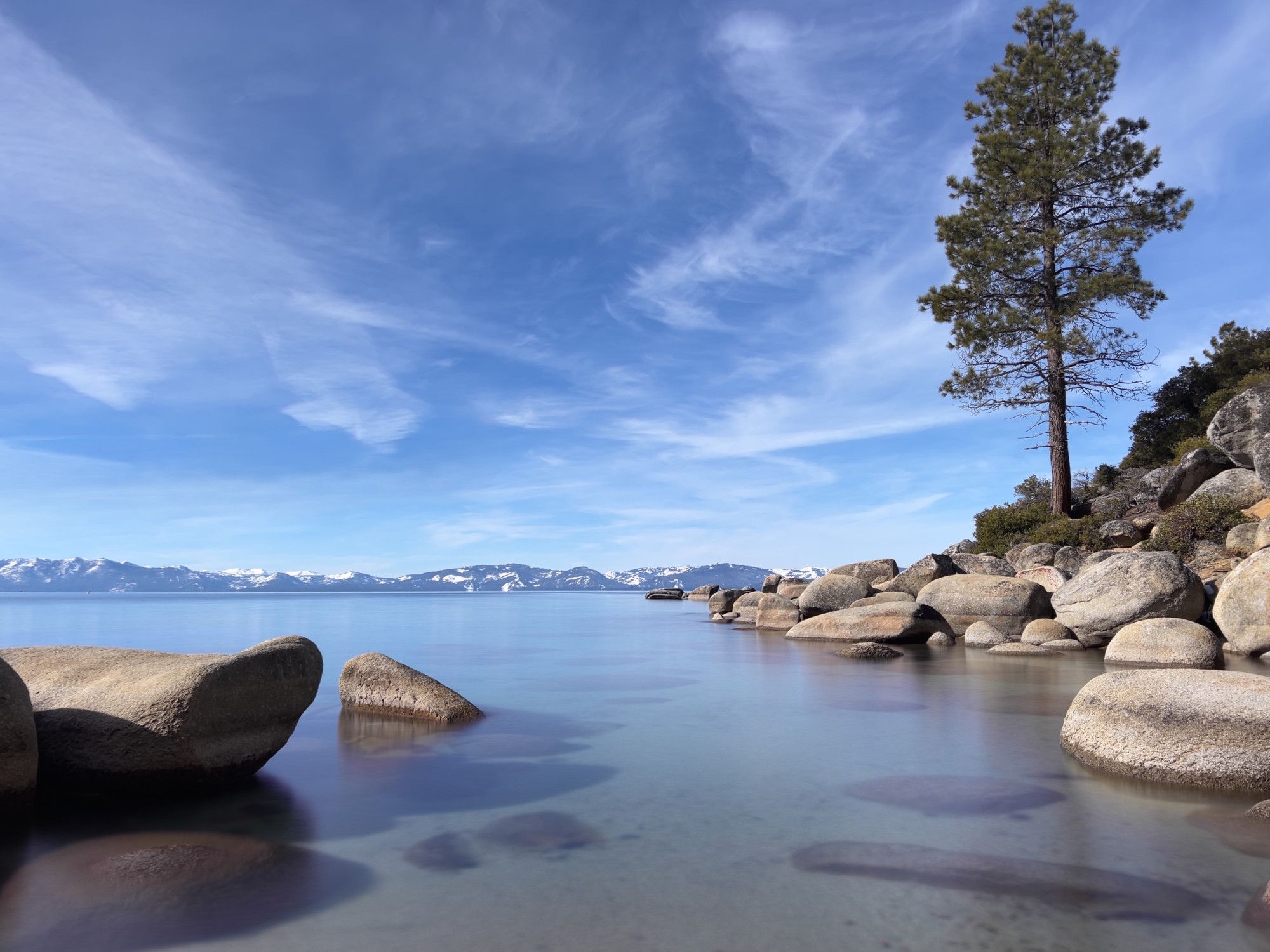 A view of a mountain lake at boulder covered shoreline, snow covered mountains in the background and a single pine tree growing from the boulders. The sky is mostly clear with some smeary clouds.