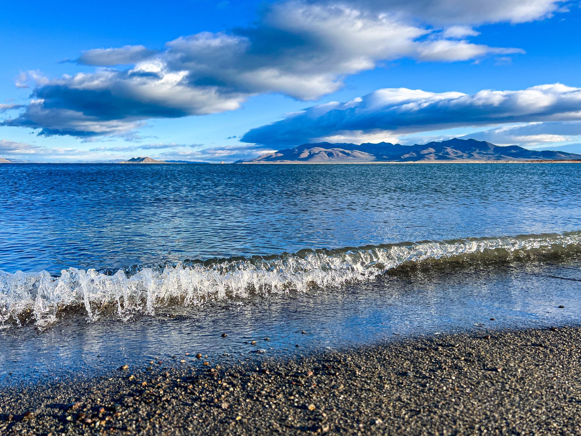 A wave crashing on a dark pebbly beach from a large desert lake. There are mountains on the horizon and big fluffy cloud in the sky.