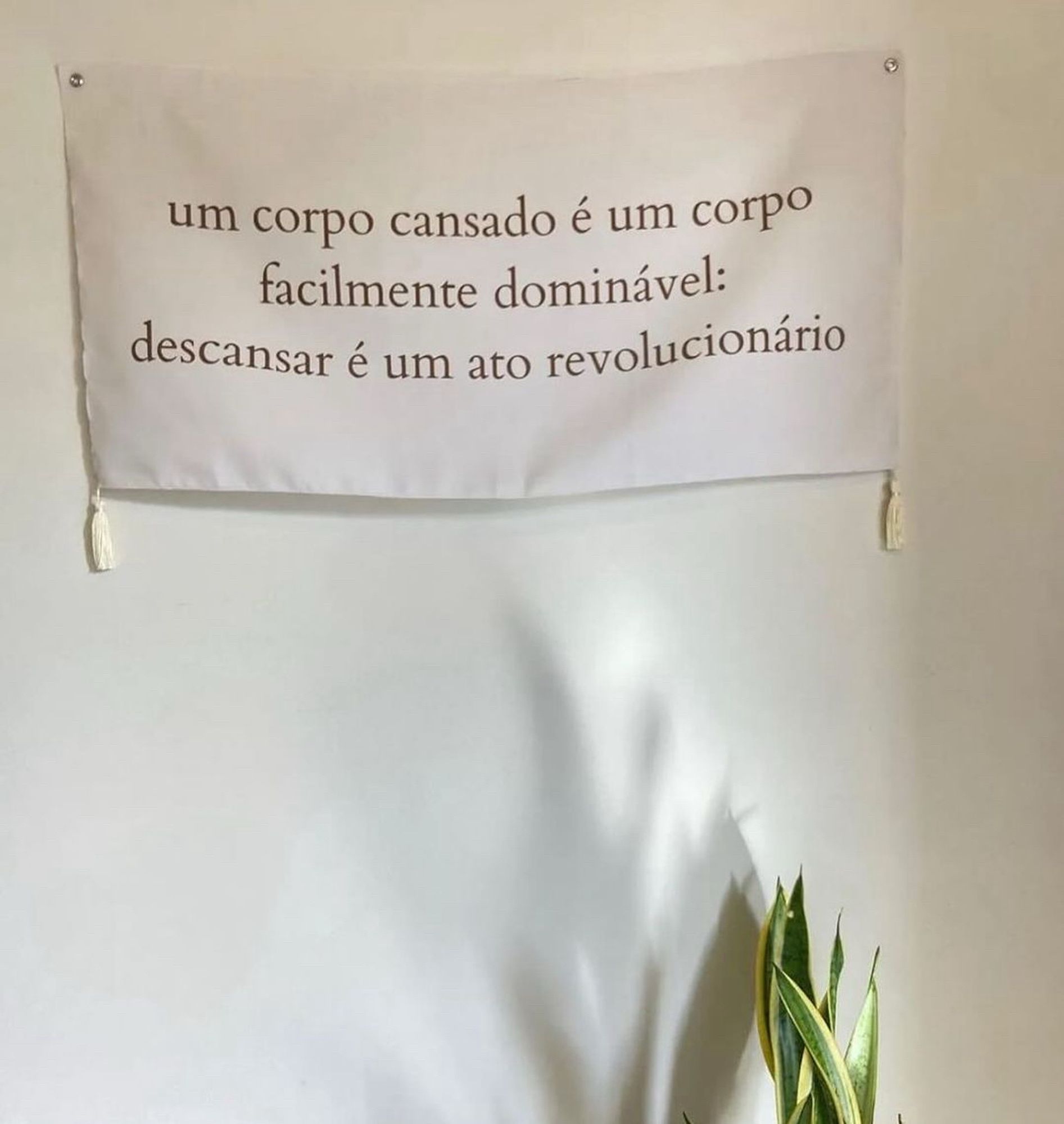 imagem de uma faixa decorativa colocada numa parede branca. a faixa diz “um corpo cansado é um corpo facilmente dominável:
descansar é um ato revolucionário”