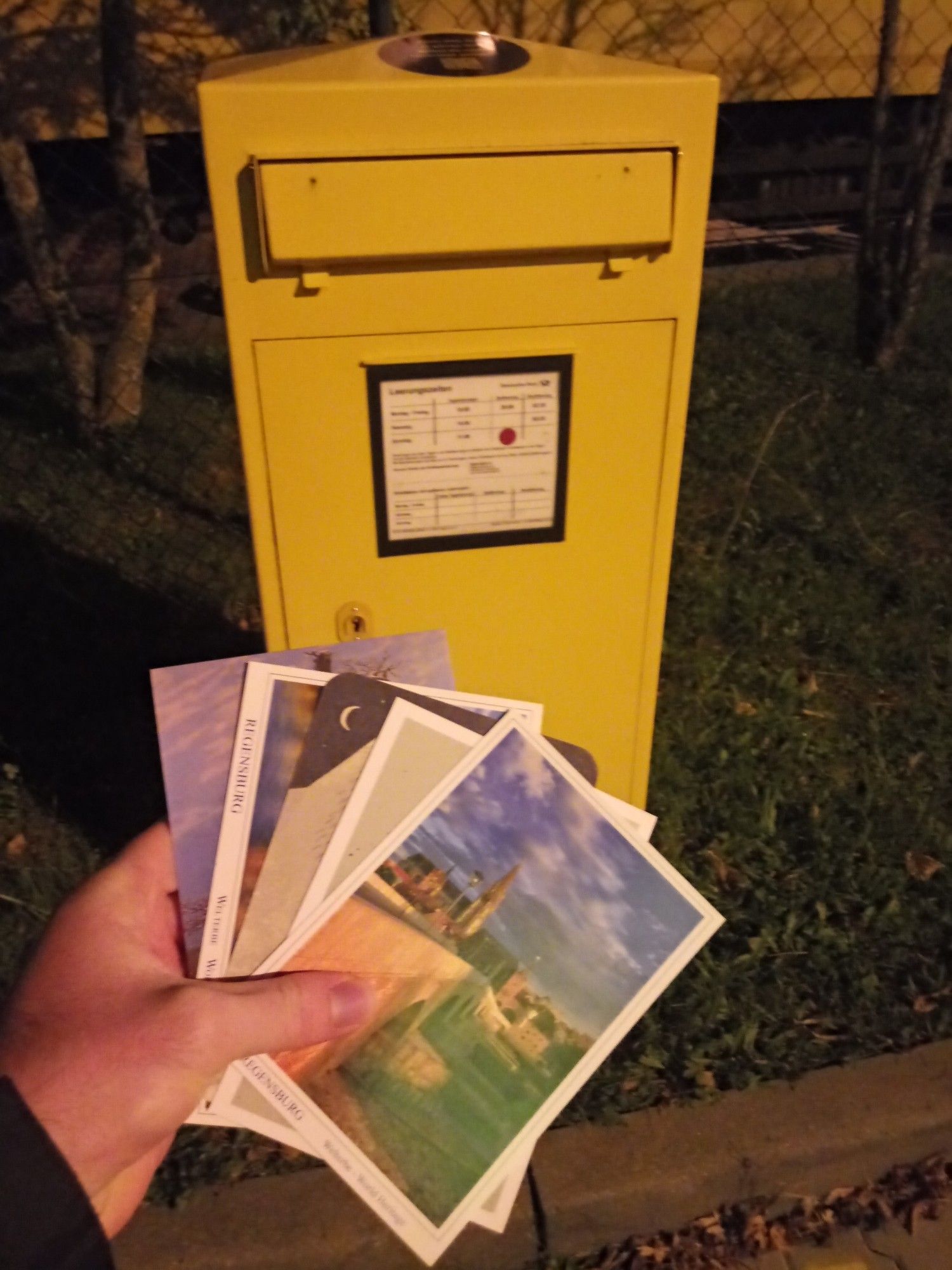 Vor einem gelben Briefkasten meine linke Hand, die fünf aufgefächerte Postkarten hält.