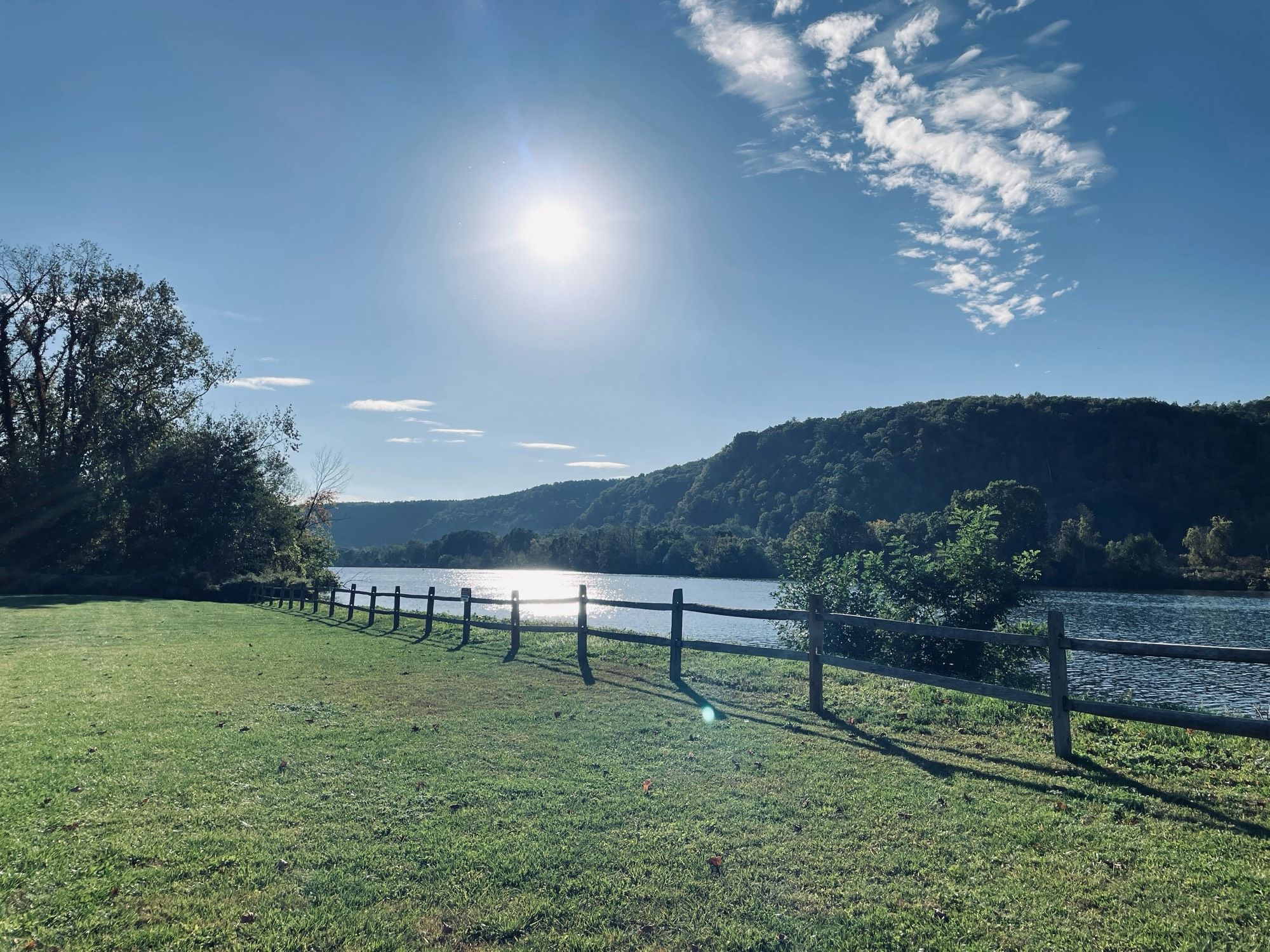 in the foreground, green grass and a low wood fence. in the background a wide calm river with a tree covered mountain. the sun is high and reflecting on the water