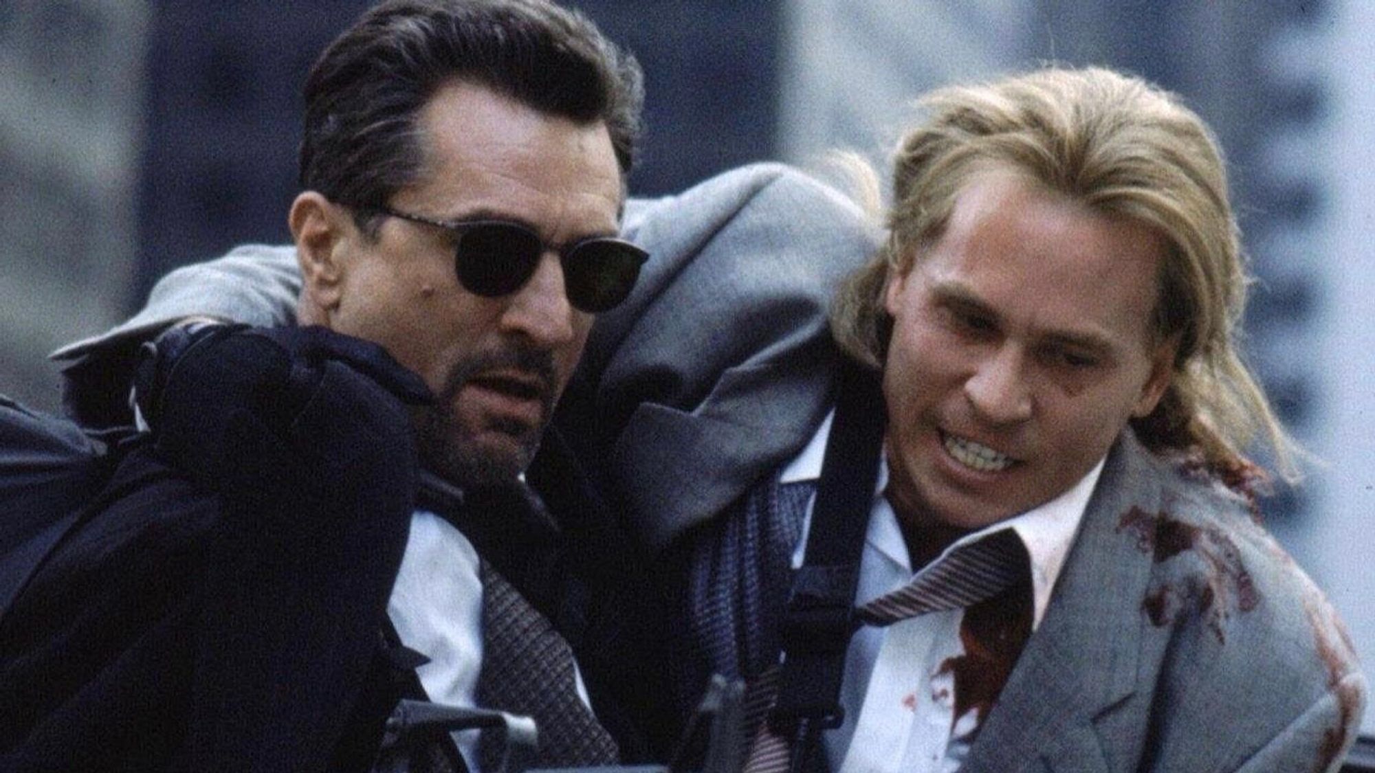 Scene from the movie Heat with long haired and bloodied Val Kilmer being propped up by Robert Deniro in shades.